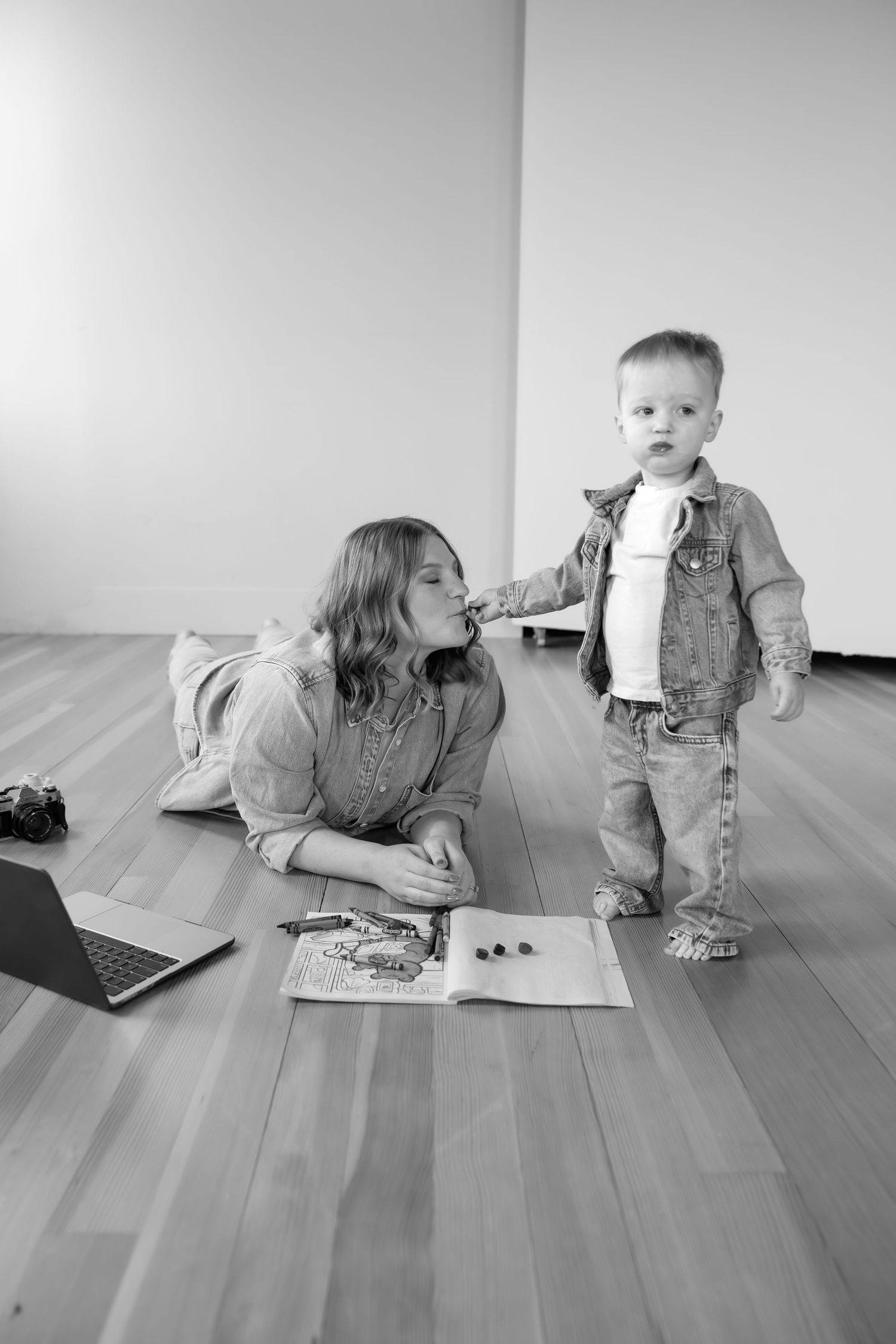 Black and white photo of a mother lying on the floor reading with her toddler son in a studio.