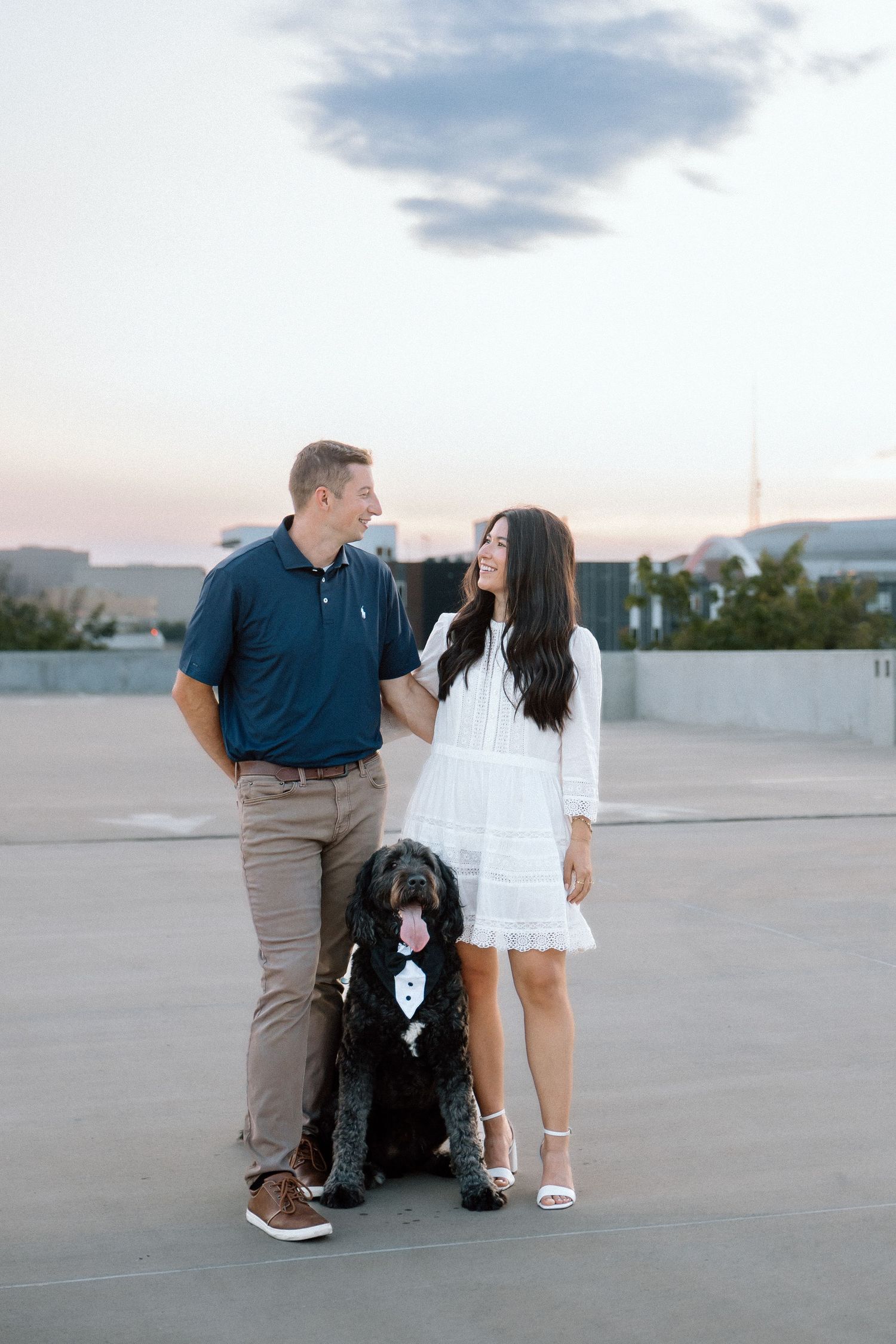 Couple smiling at each other with their black doodle dog in a tuxedo on a rooftop at sunset.