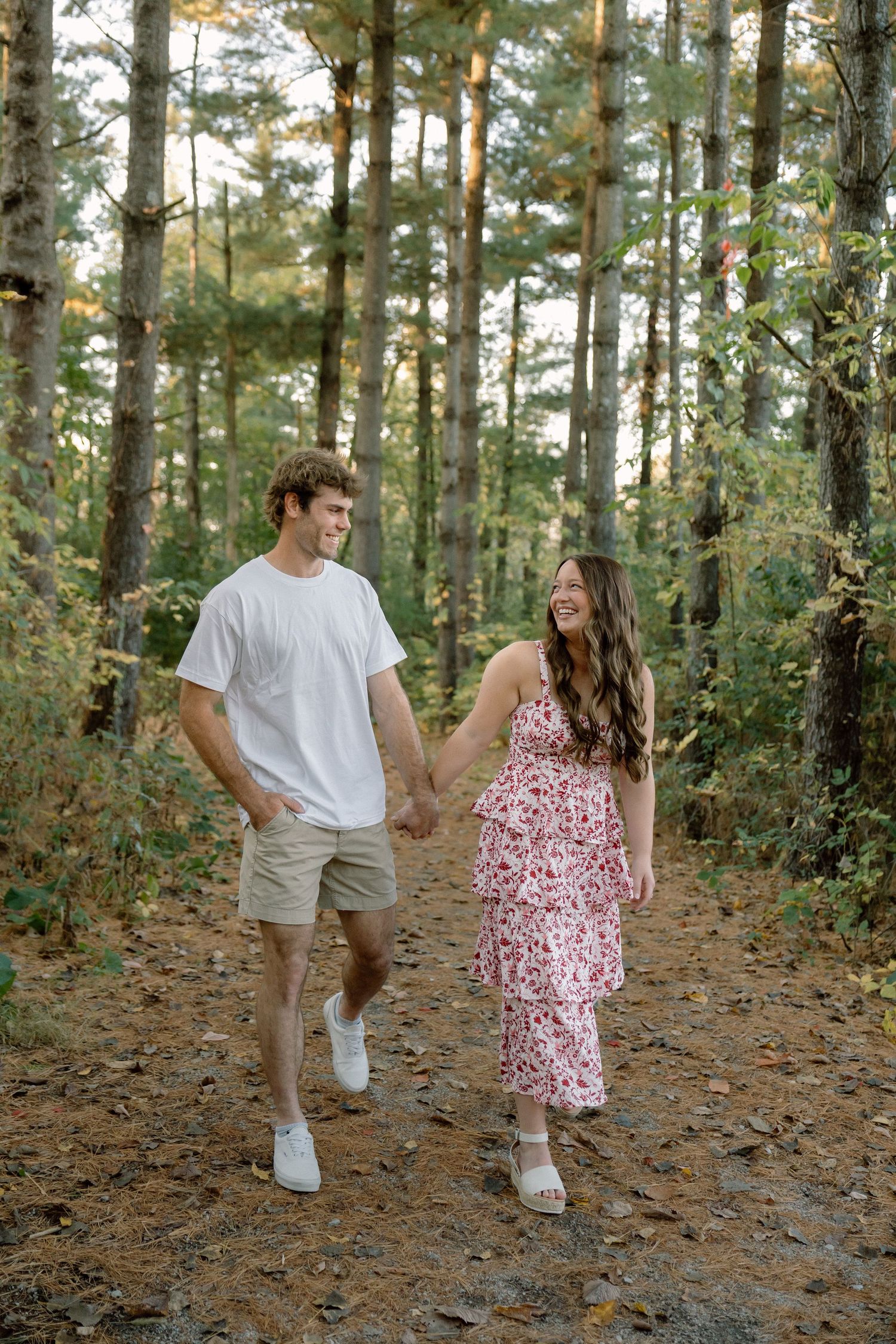 Couple holding hands walking on a forest trail, woman in floral dress and man in white tee and khaki shorts.