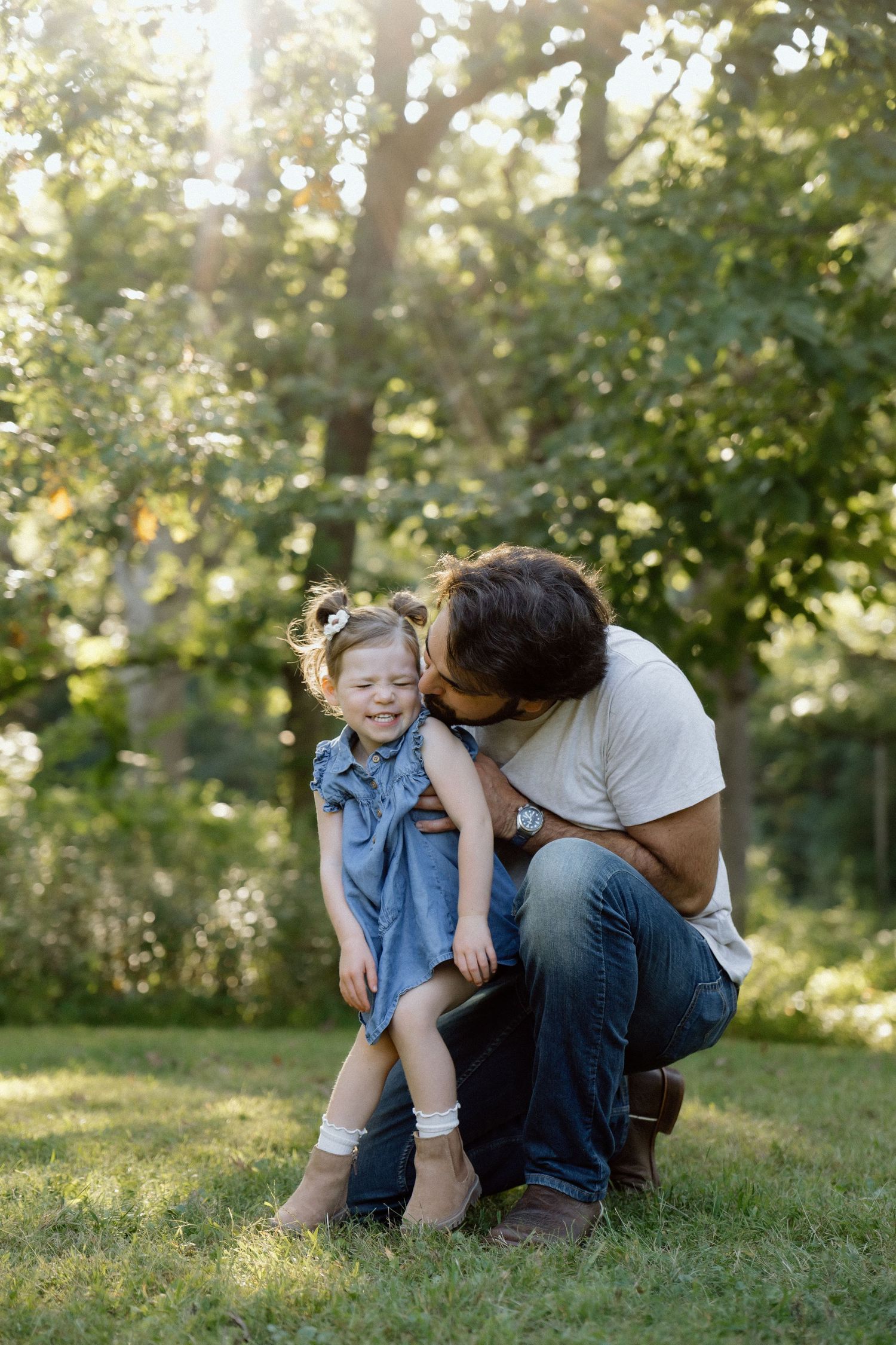 Loving father kneeling to kiss smiling young daughter in blue dress in sunlit park.