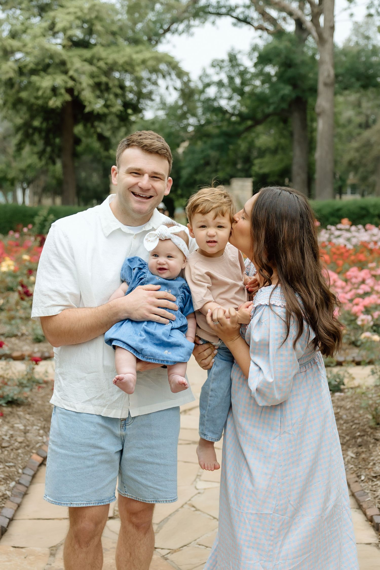 Smiling family of four poses in rose garden, father holding infant in denim while mother kisses toddler.