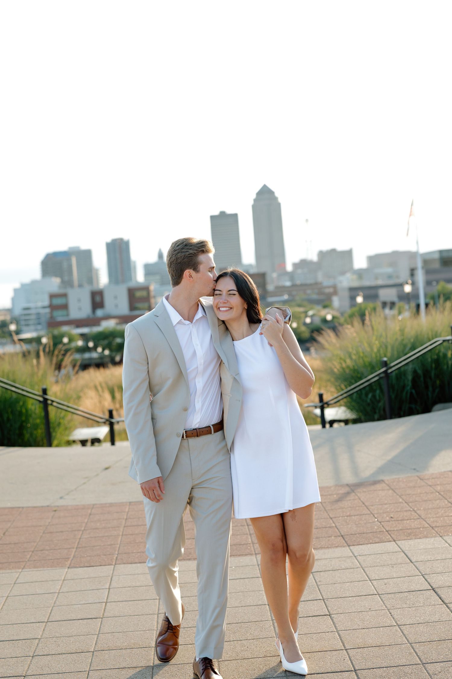 Couple in elegant attire sharing a romantic moment with city skyline backdrop, man in tan suit kissing woman in white dress.
