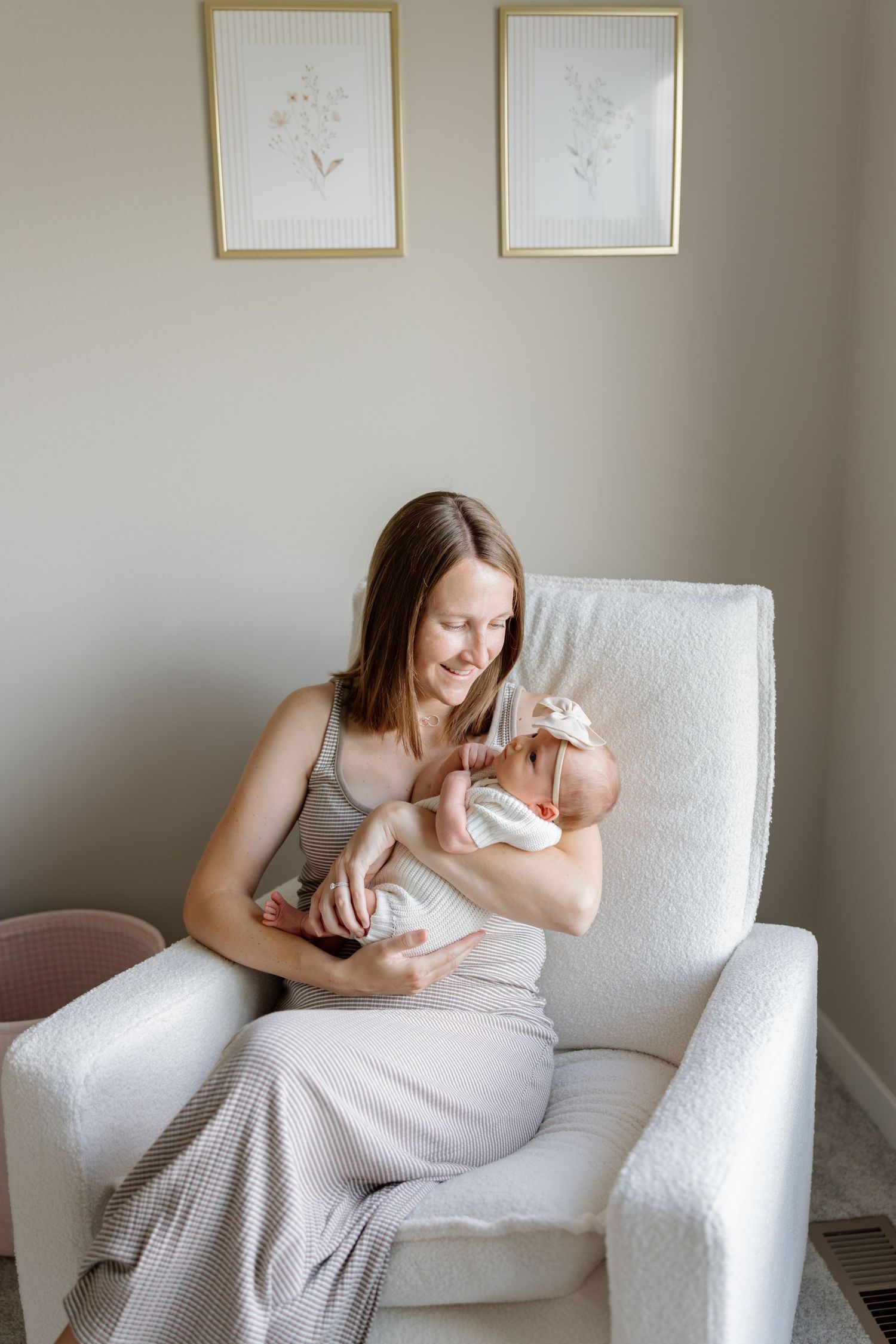 A smiling mother sits in a white glider chair holding her newborn baby in a softly decorated nursery.