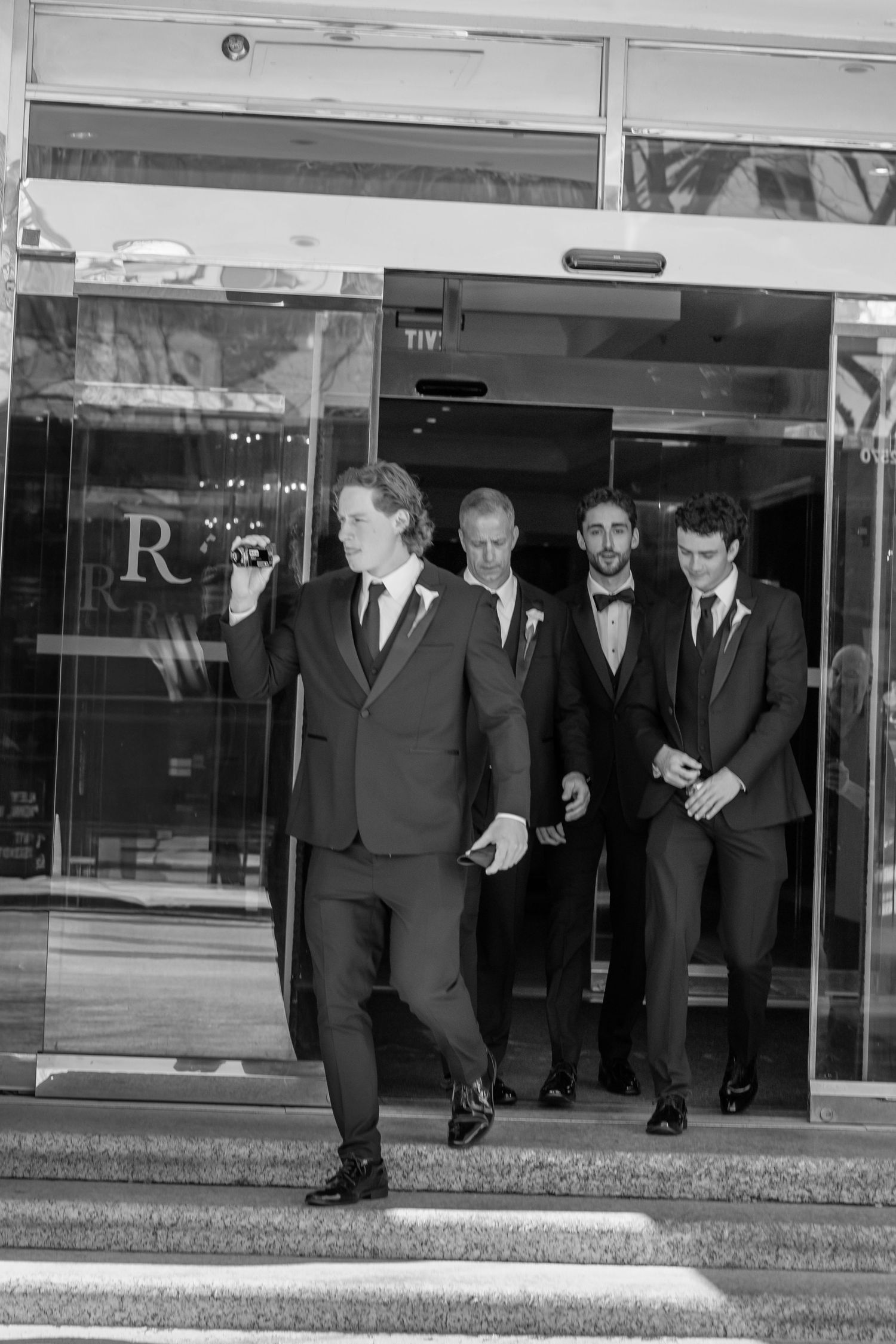 Groomsmen in black suits exit through glass doors of an elegant venue on a wedding day.