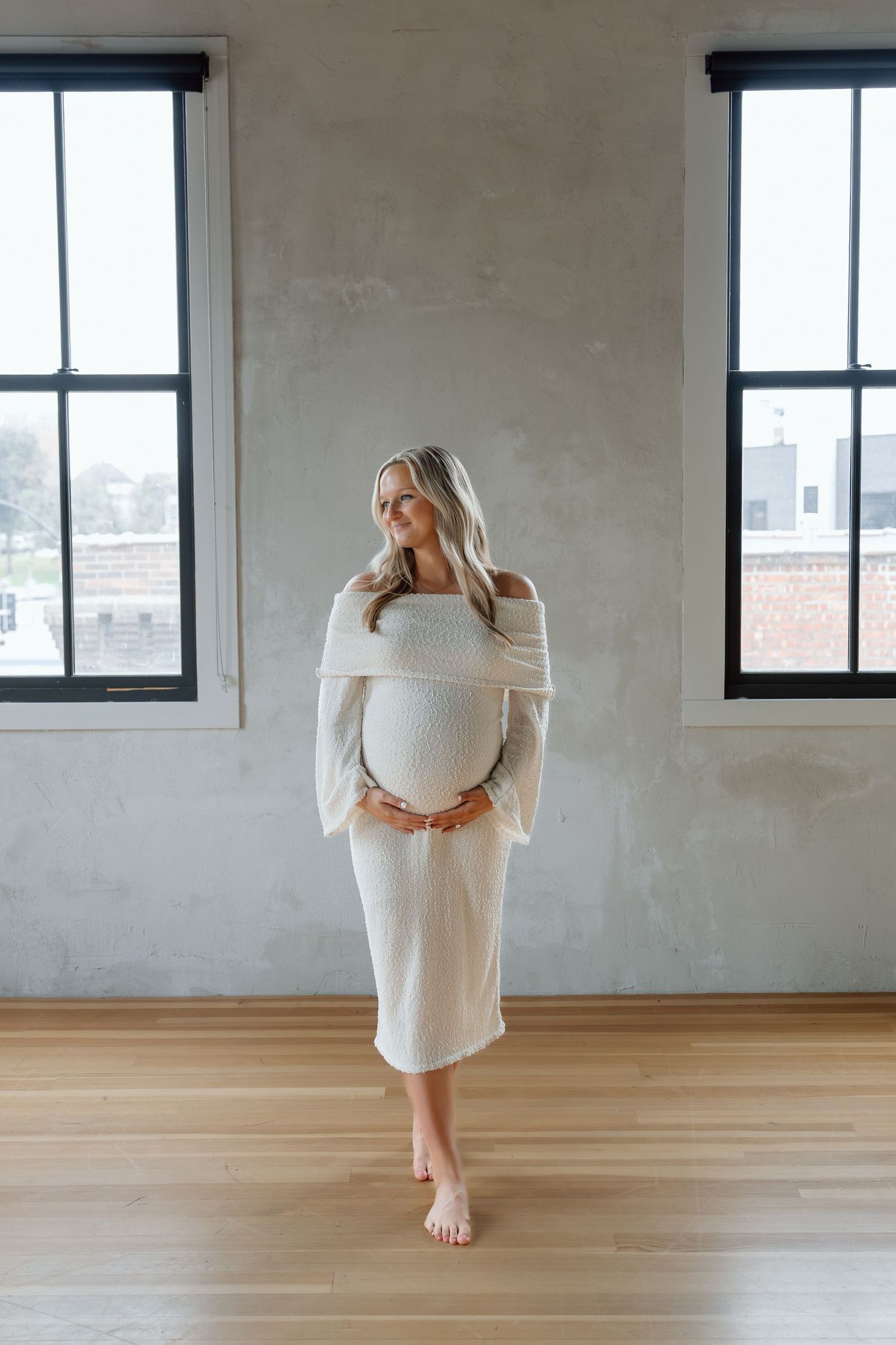 Pregnant woman in cream off-shoulder knit dress holding baby bump near large windows.