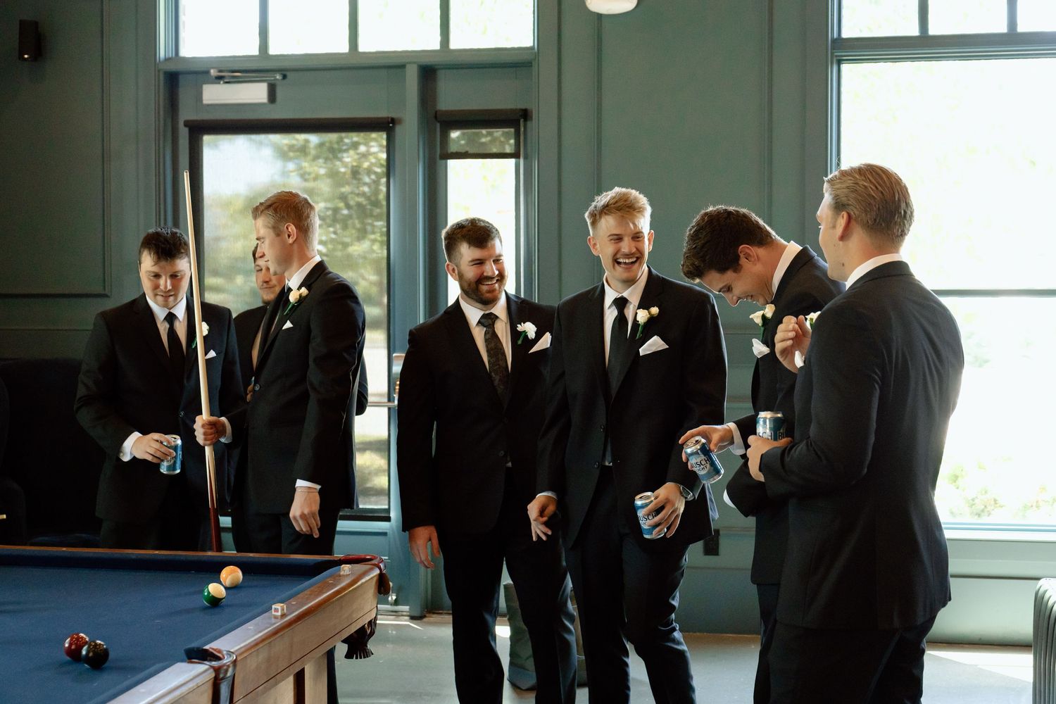 Groomsmen in black suits laughing together near a pool table before a wedding ceremony at The Monroe wedding venue in West Des Moines, Iowa