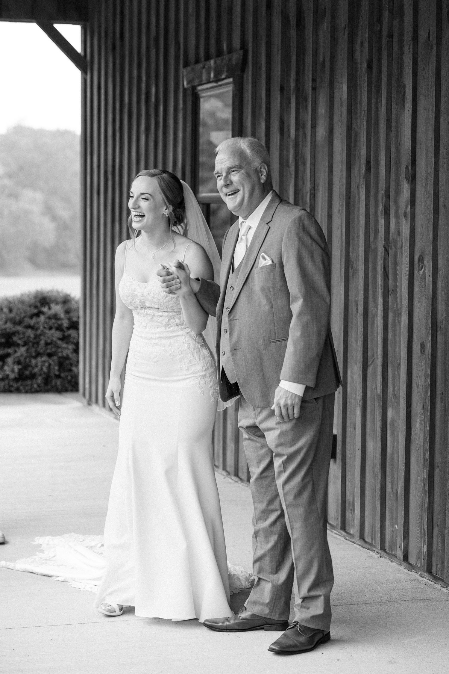 Black and white photo of a smiling bride in a lace gown walking arm-in-arm with her father at a barn wedding.