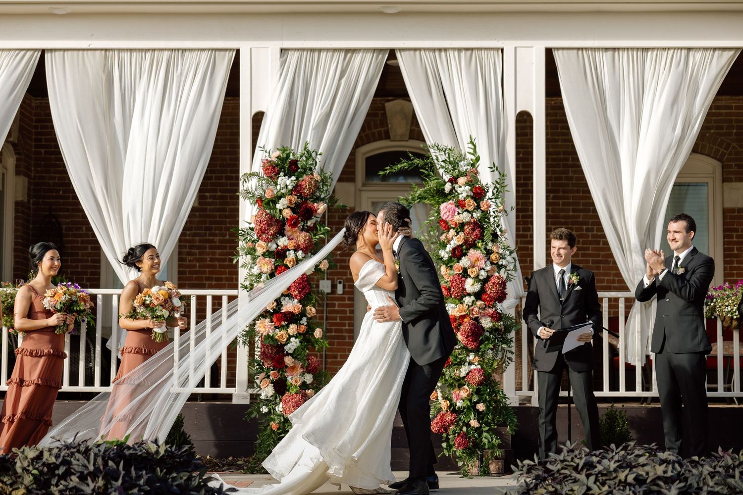 Bride and groom share first kiss at altar adorned with red and white floral arrangements on venue porch.