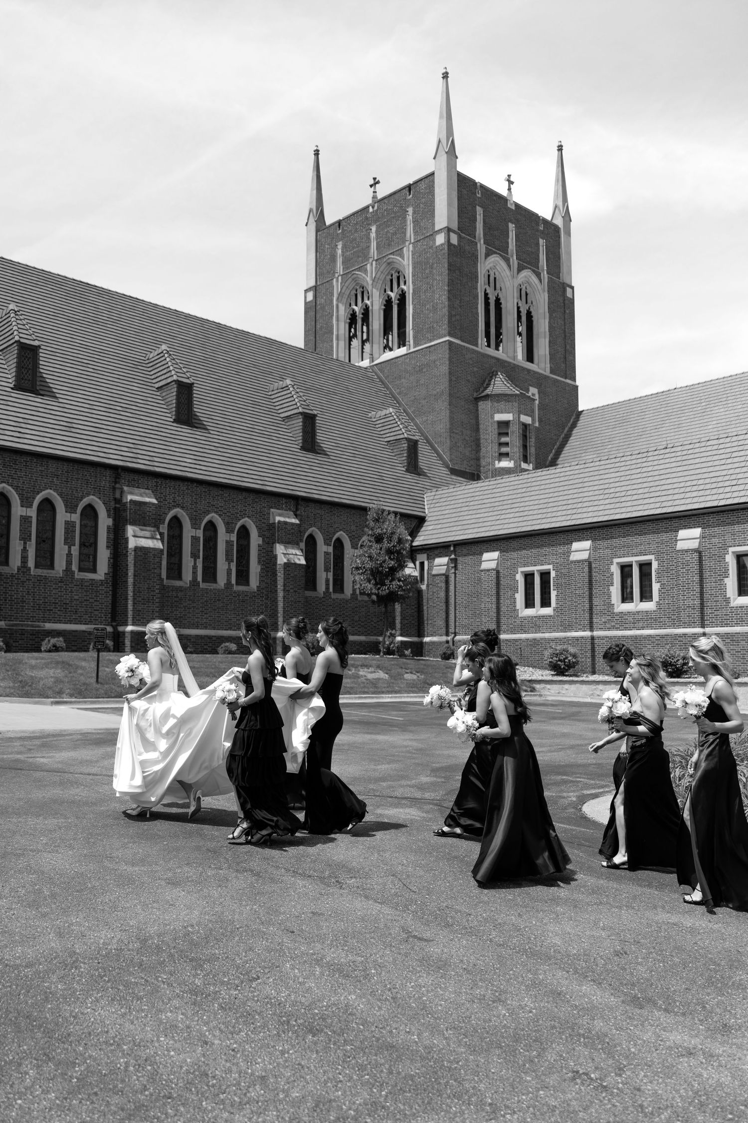 Black and white photo of a bride and bridesmaids in dark gowns walking outside a brick church.