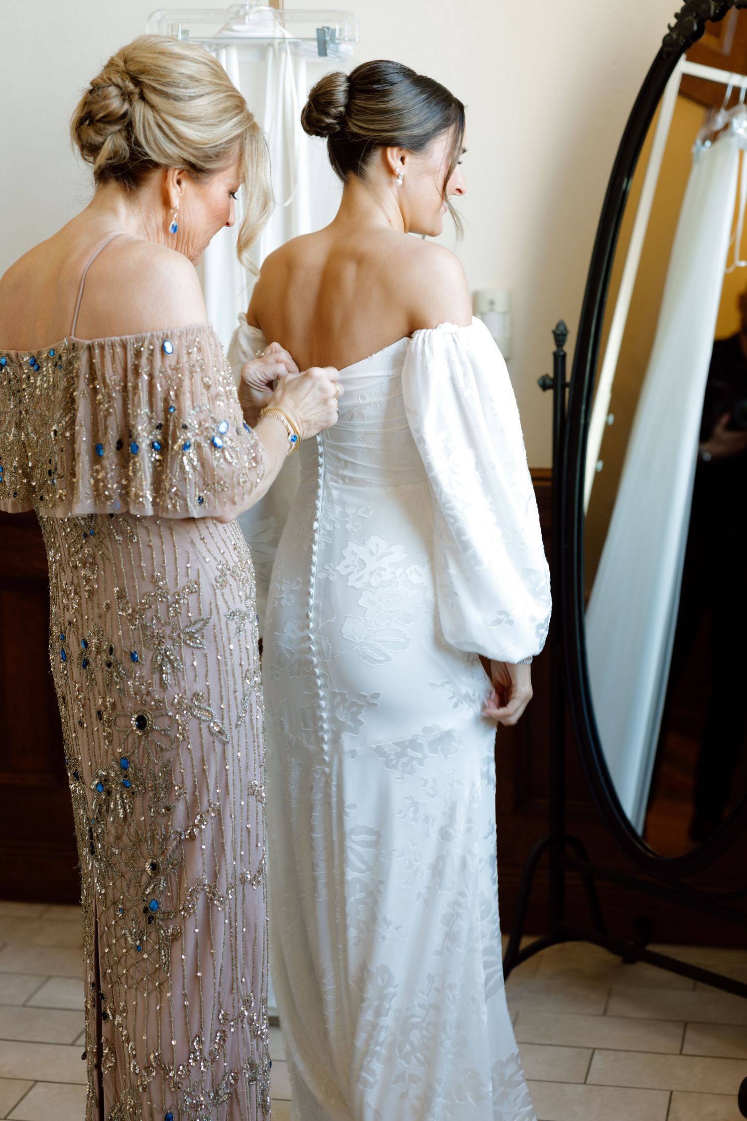 Bridesmaid in beaded gown buttons the back of a brides white off-shoulder wedding dress.