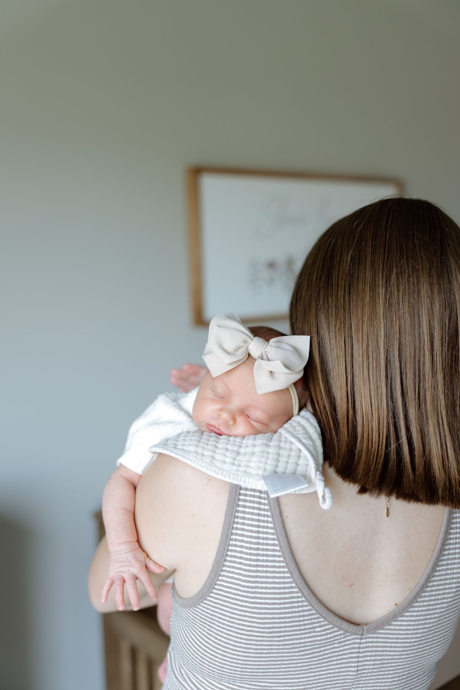 Mother holding sleeping newborn baby girl wearing a white bow headband over her shoulder in a nursery.