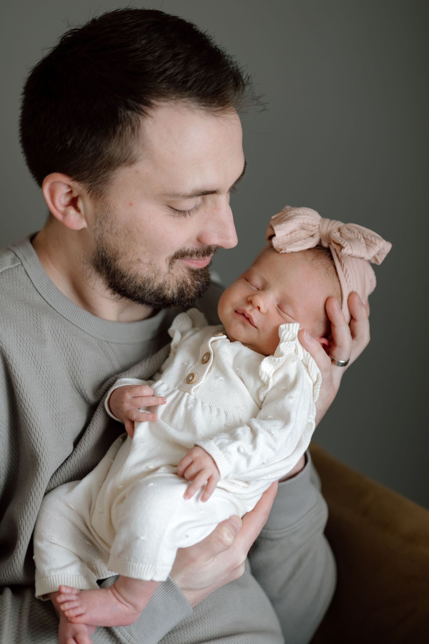 Loving father in grey shirt smiling at sleeping newborn daughter with pink bow.