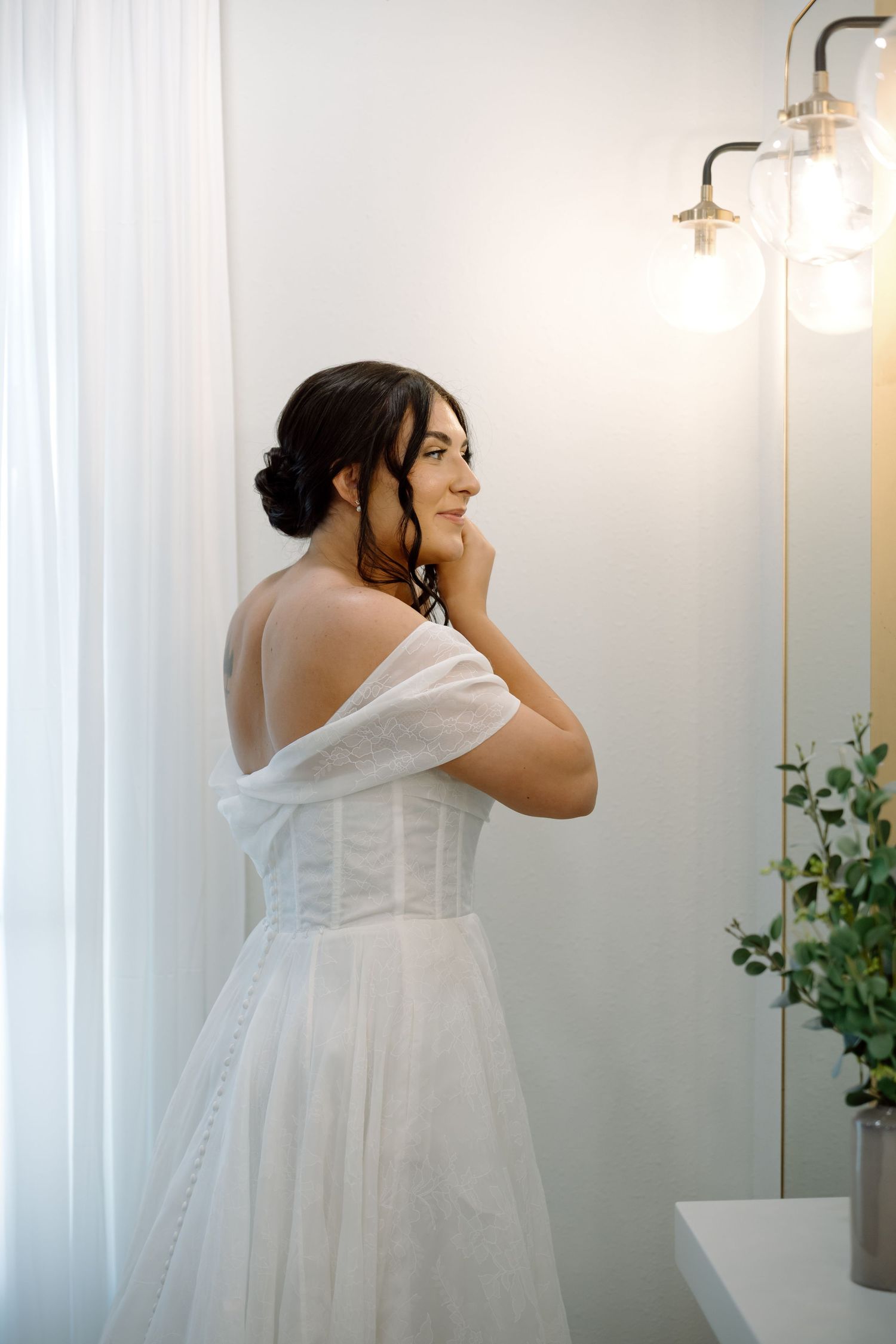 Elegant bride in off-shoulder corset wedding gown touching her ear while standing near a softly lit mirror.