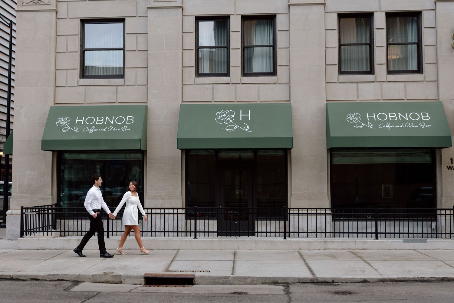 Couple holding hands walking past Hobnob restaurant with green awnings on a city street.