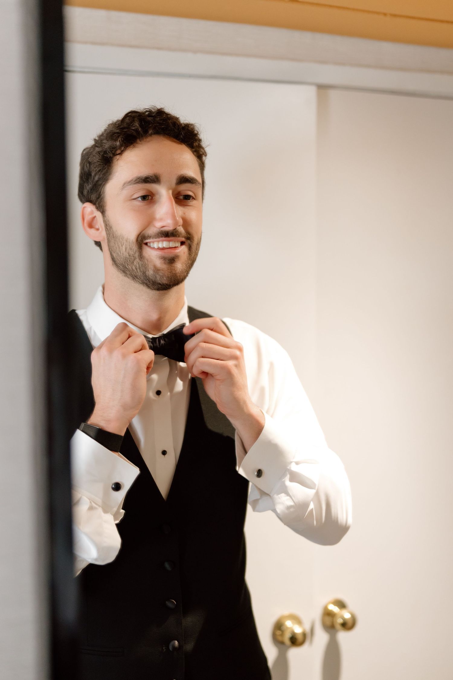 Smiling groom adjusting black bow tie wearing white tuxedo shirt and black vest while getting ready.