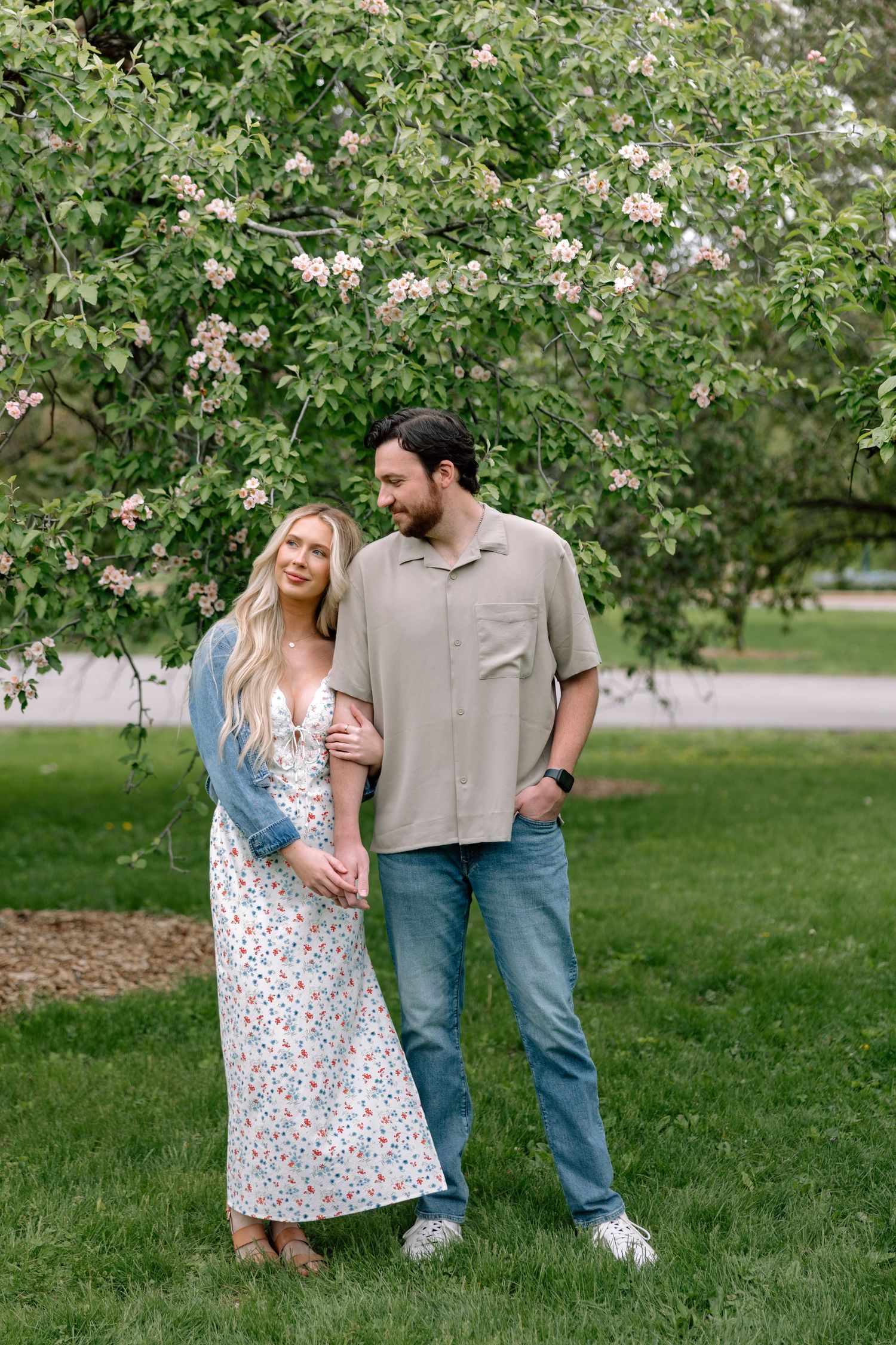A couple stands together under a blooming tree in a lush green park, smiling warmly on a spring day.