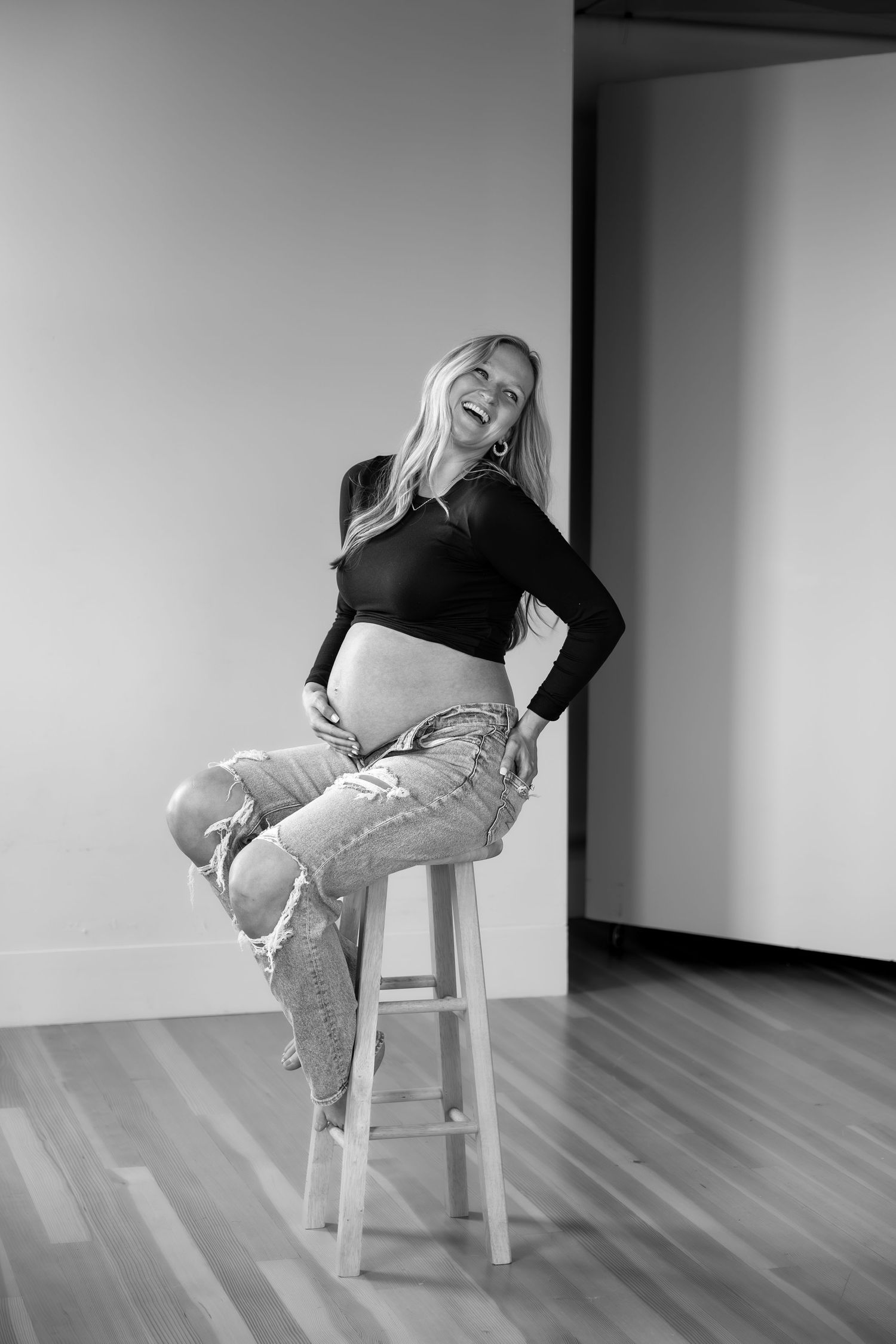 Black and white photo of laughing pregnant woman sitting on stool in ripped jeans and crop top.