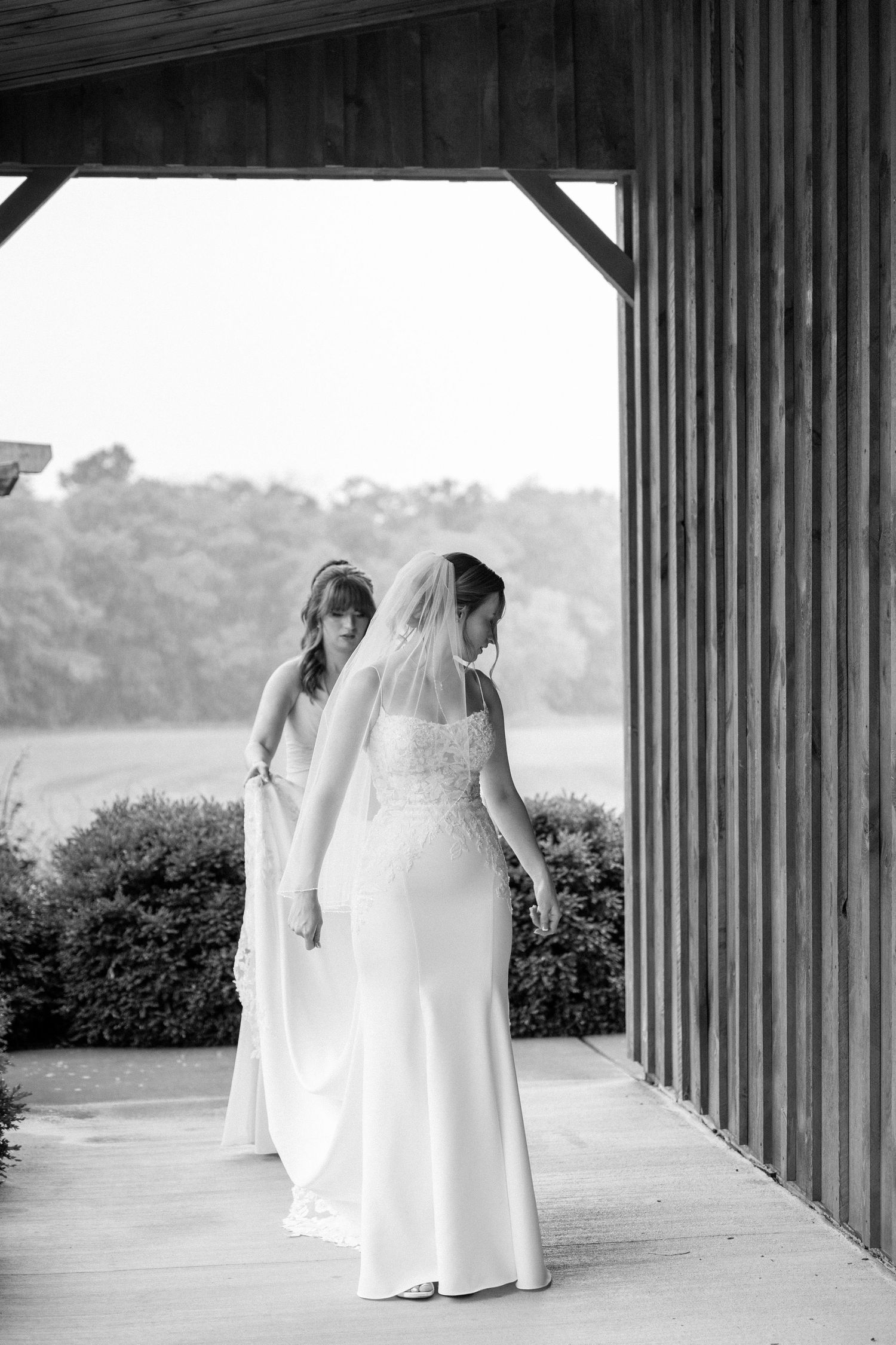 Black and white photo of a bridesmaid adjusting the brides veil outside a barn venue.