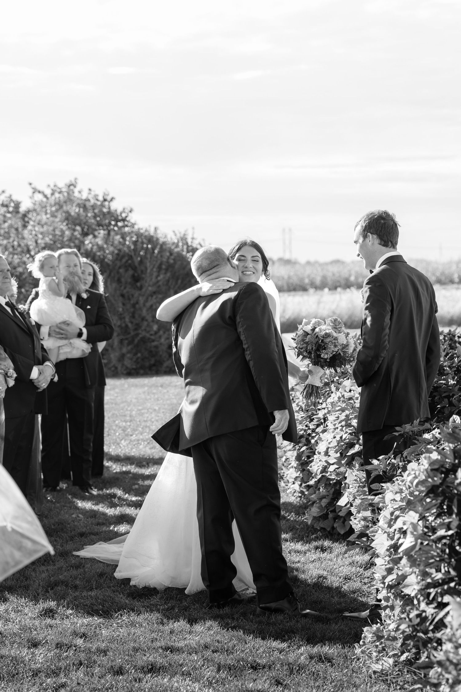 Black and white photo of bride embracing her father at outdoor wedding ceremony while groom watches nearby.