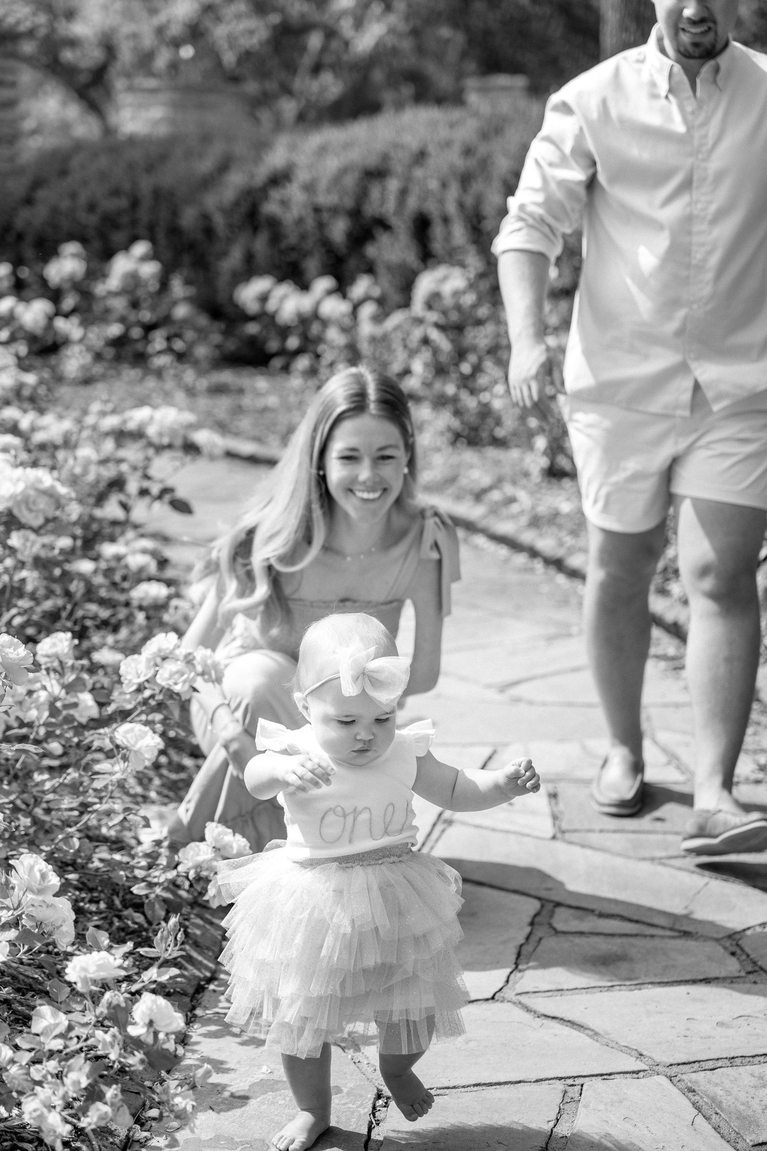 Black and white photo of smiling mother guiding toddler girl in tutu dress along garden stone path.