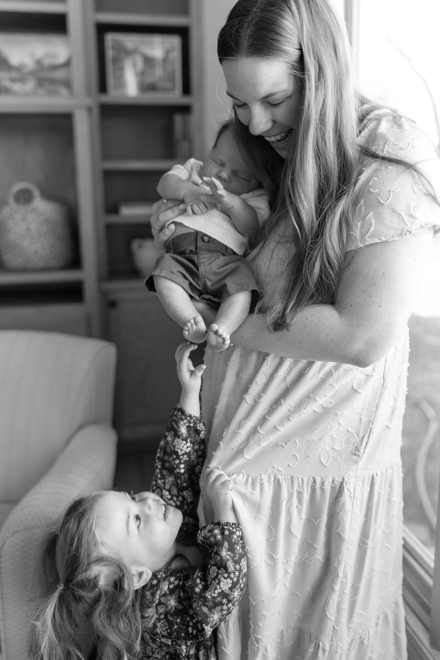 Black and white photo of a mother holding newborn as toddler reaches up.