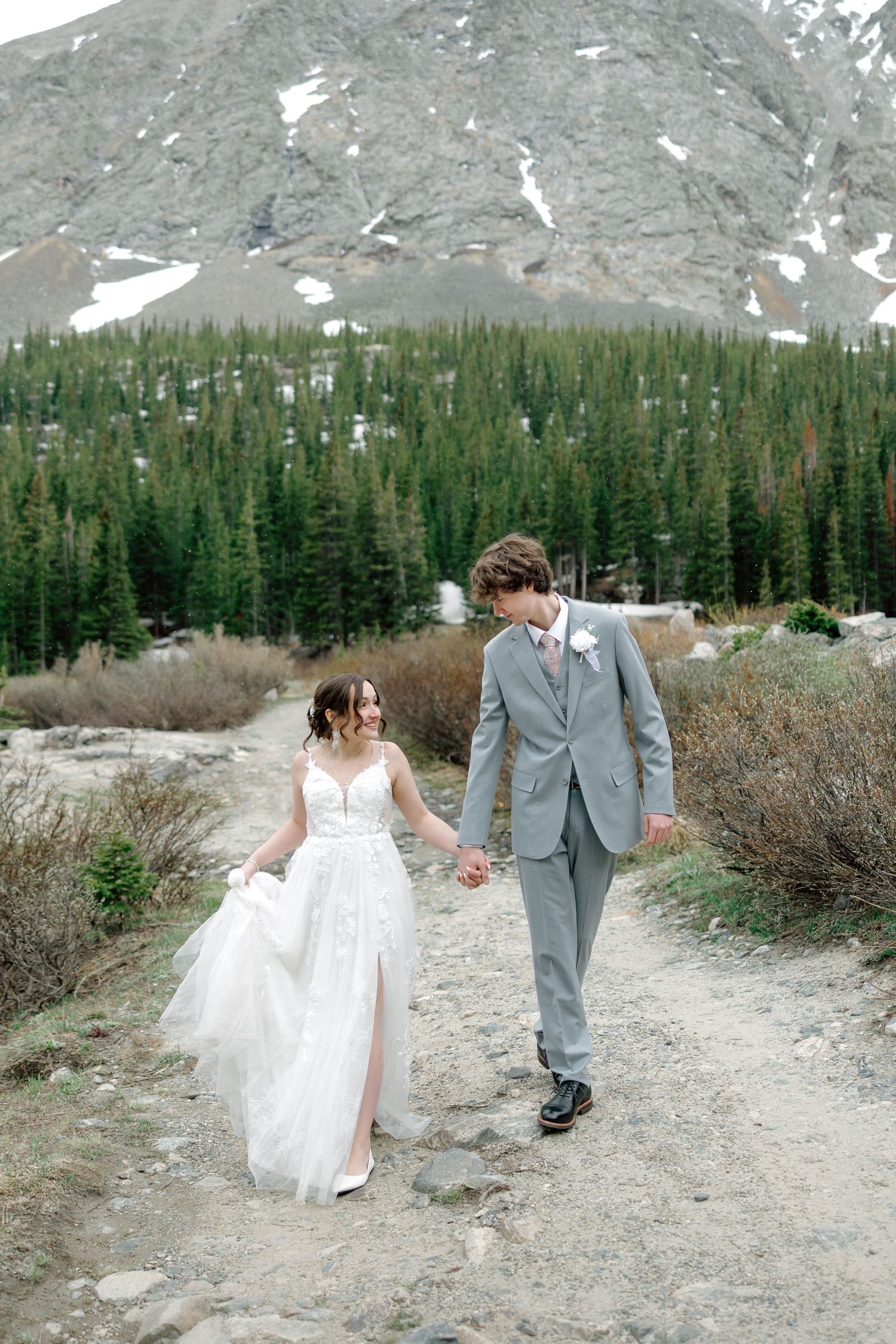 A bride and groom walk hand-in-hand on a mountain trail surrounded by pine trees and snow-capped peaks.