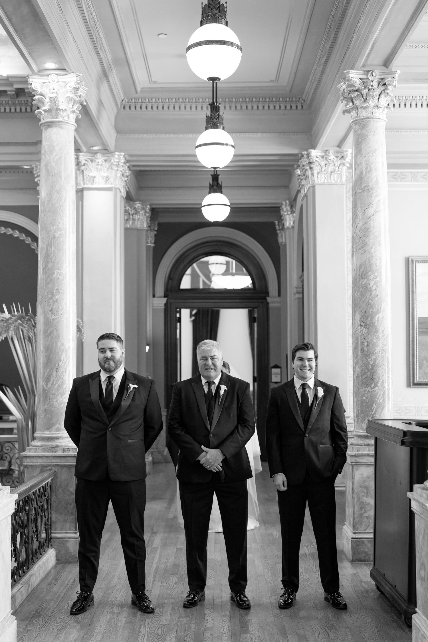 Three groomsmen in black suits stand beneath chandeliers in a grand columned wedding venue.