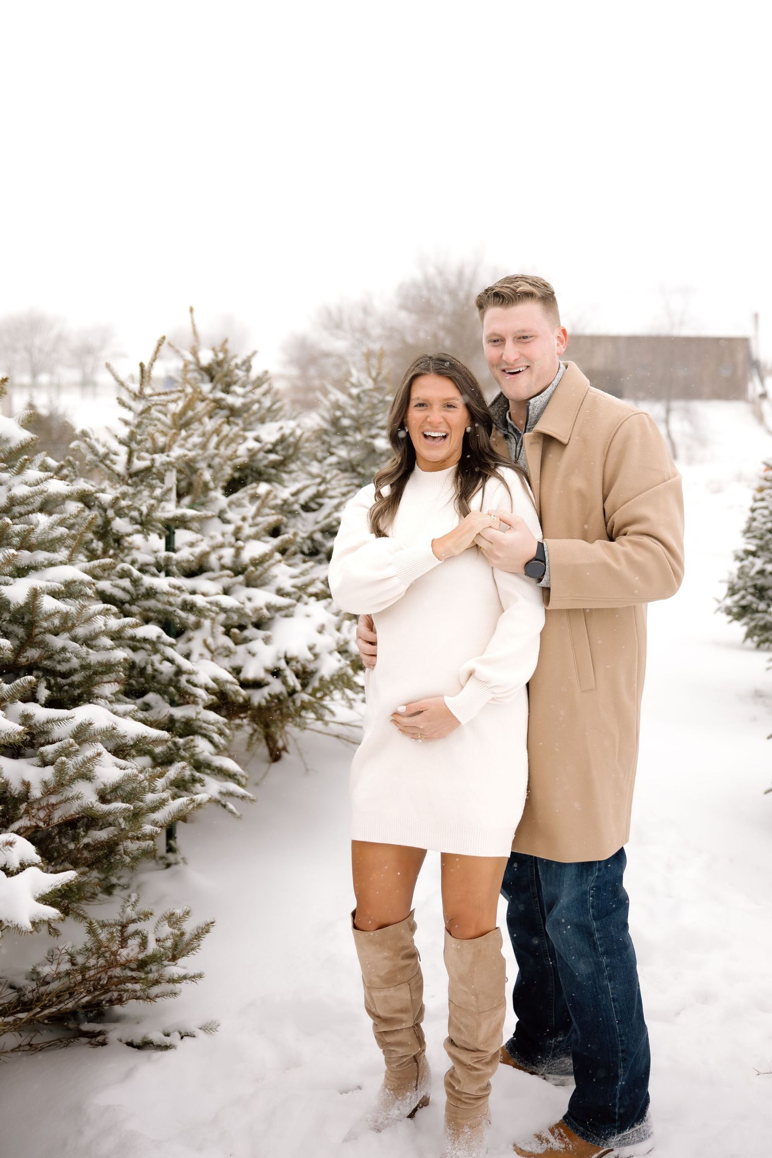 Smiling couple embracing at snowy tree farm, pregnant woman in white dress and knee-high boots.