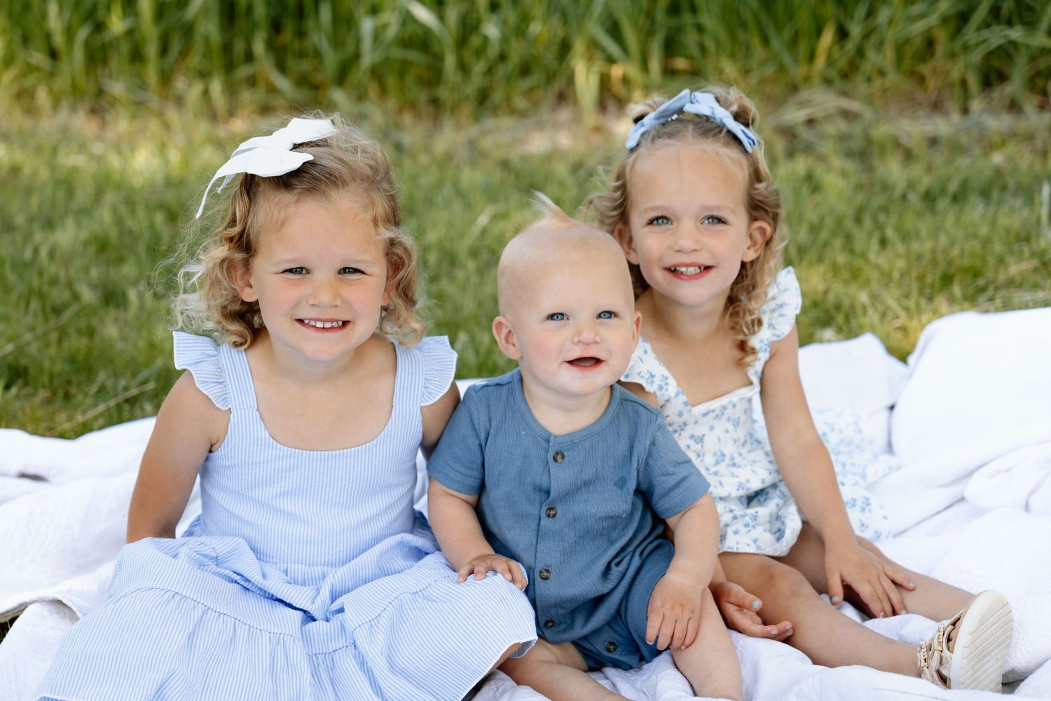 Three young siblings, two girls and a baby boy, sitting together on a white blanket outdoors.