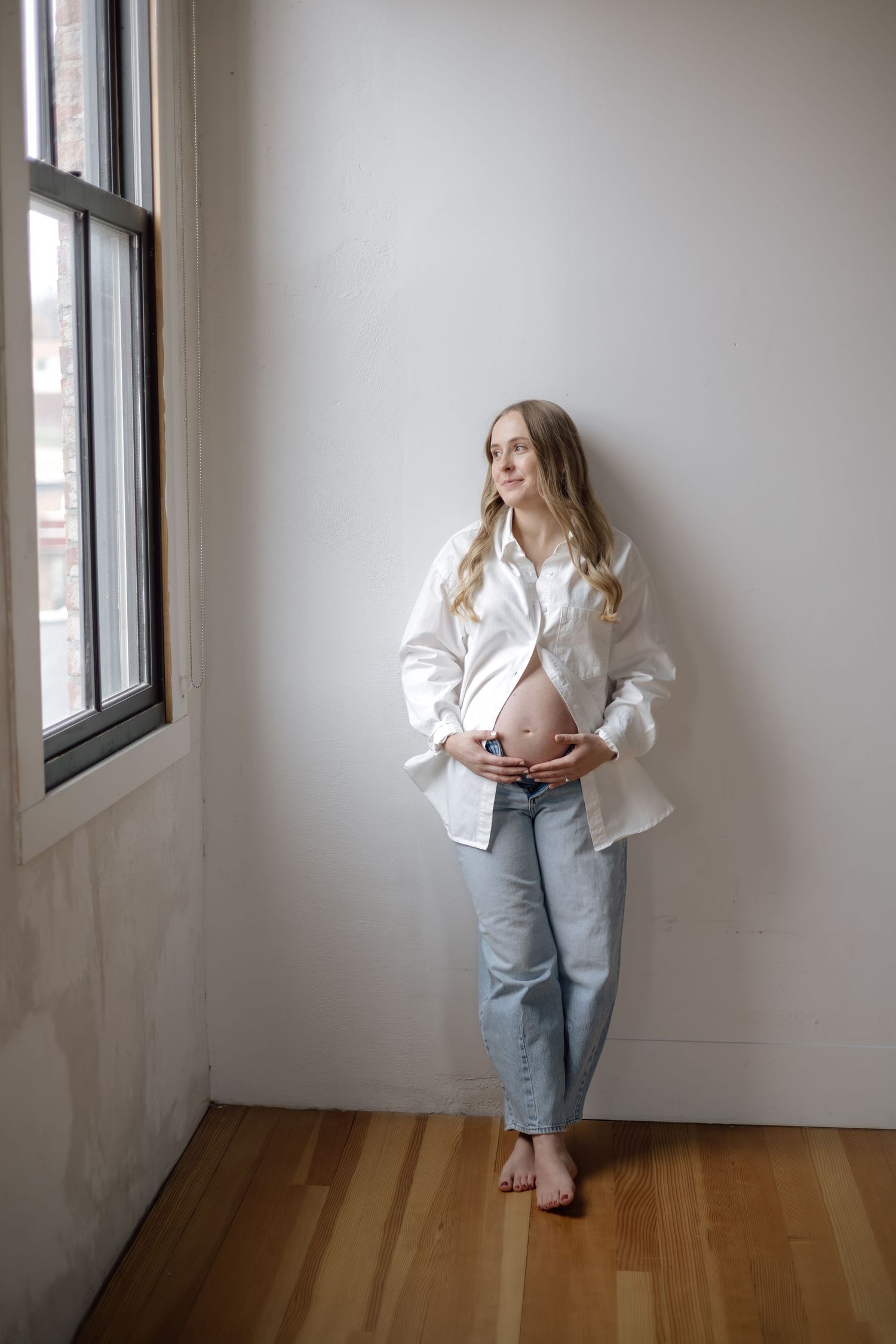 A pregnant woman in a white shirt and jeans stands by a window cradling her bare baby bump.