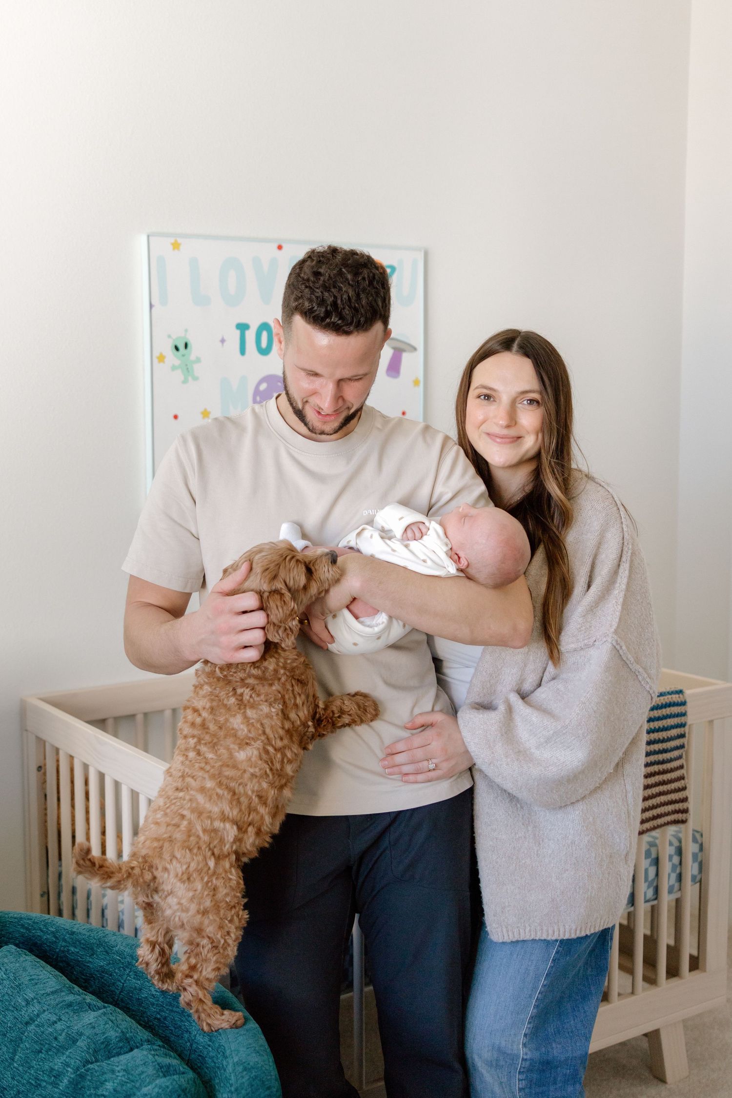 Smiling new parents hold their newborn while their fluffy brown doodle dog stands up curiously in the nursery.