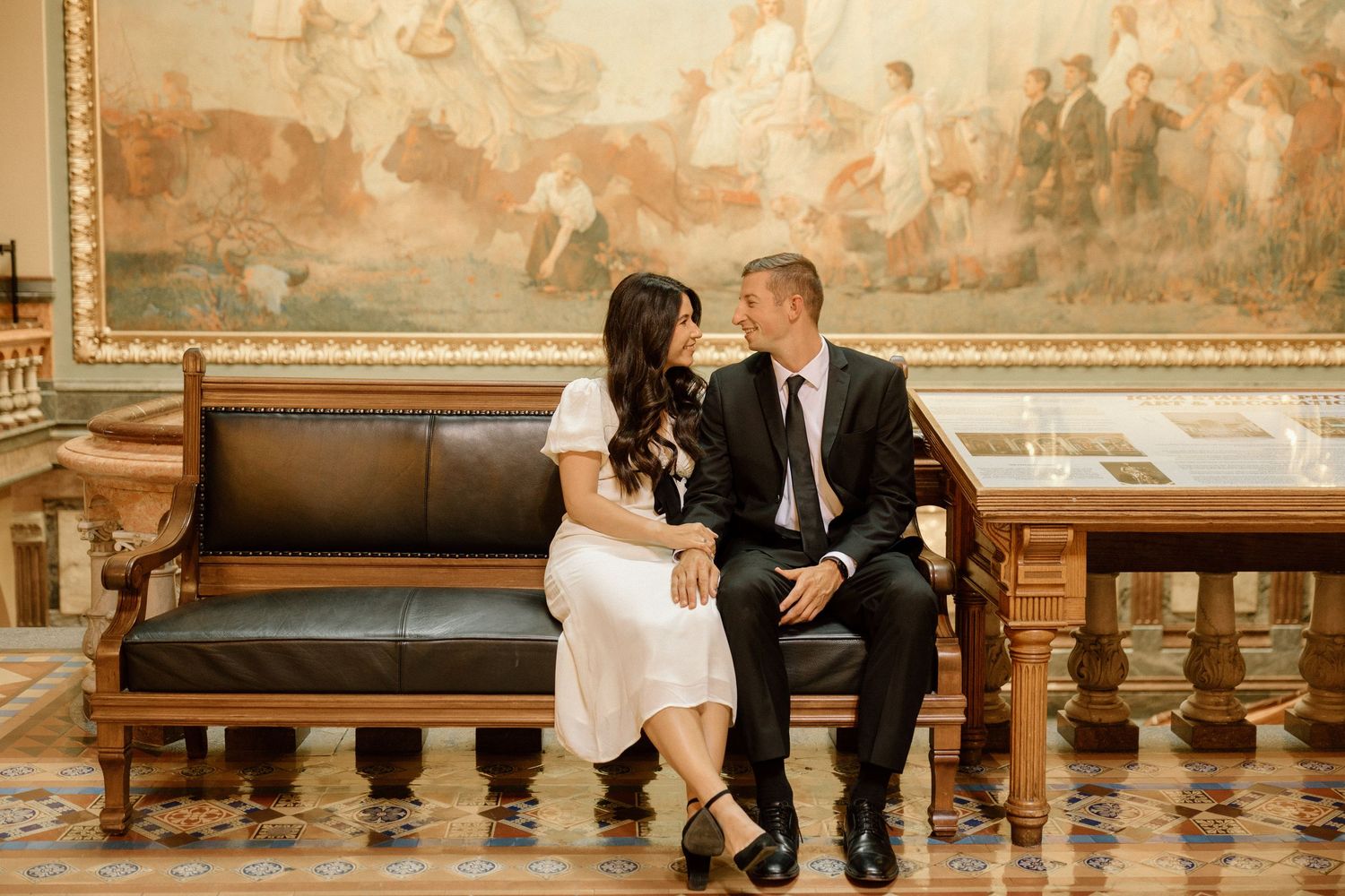 Couple sitting closely on a leather bench in an ornate hall with a large classical mural.