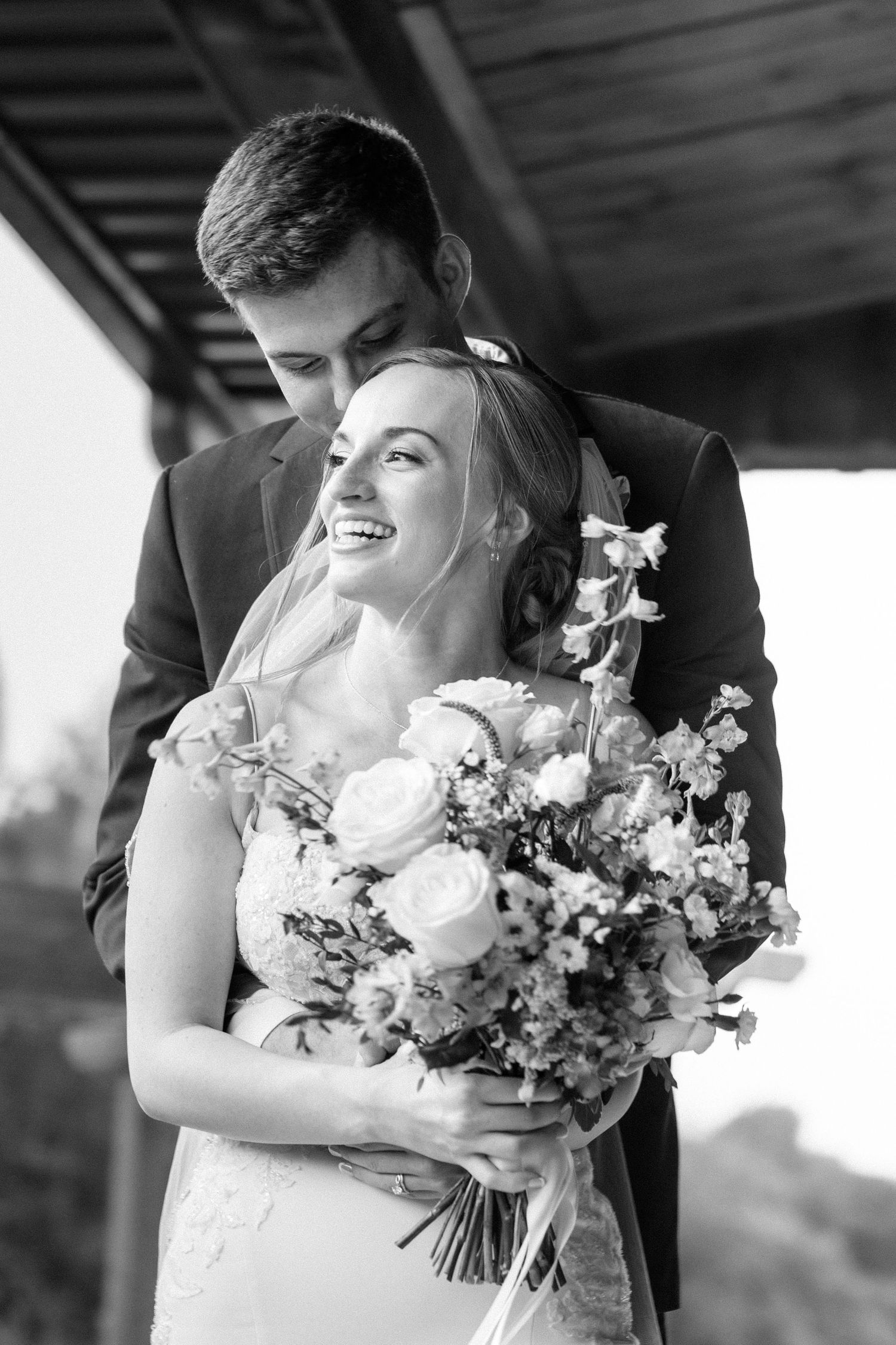 Black and white portrait of a smiling bride holding a floral bouquet while the groom embraces her.