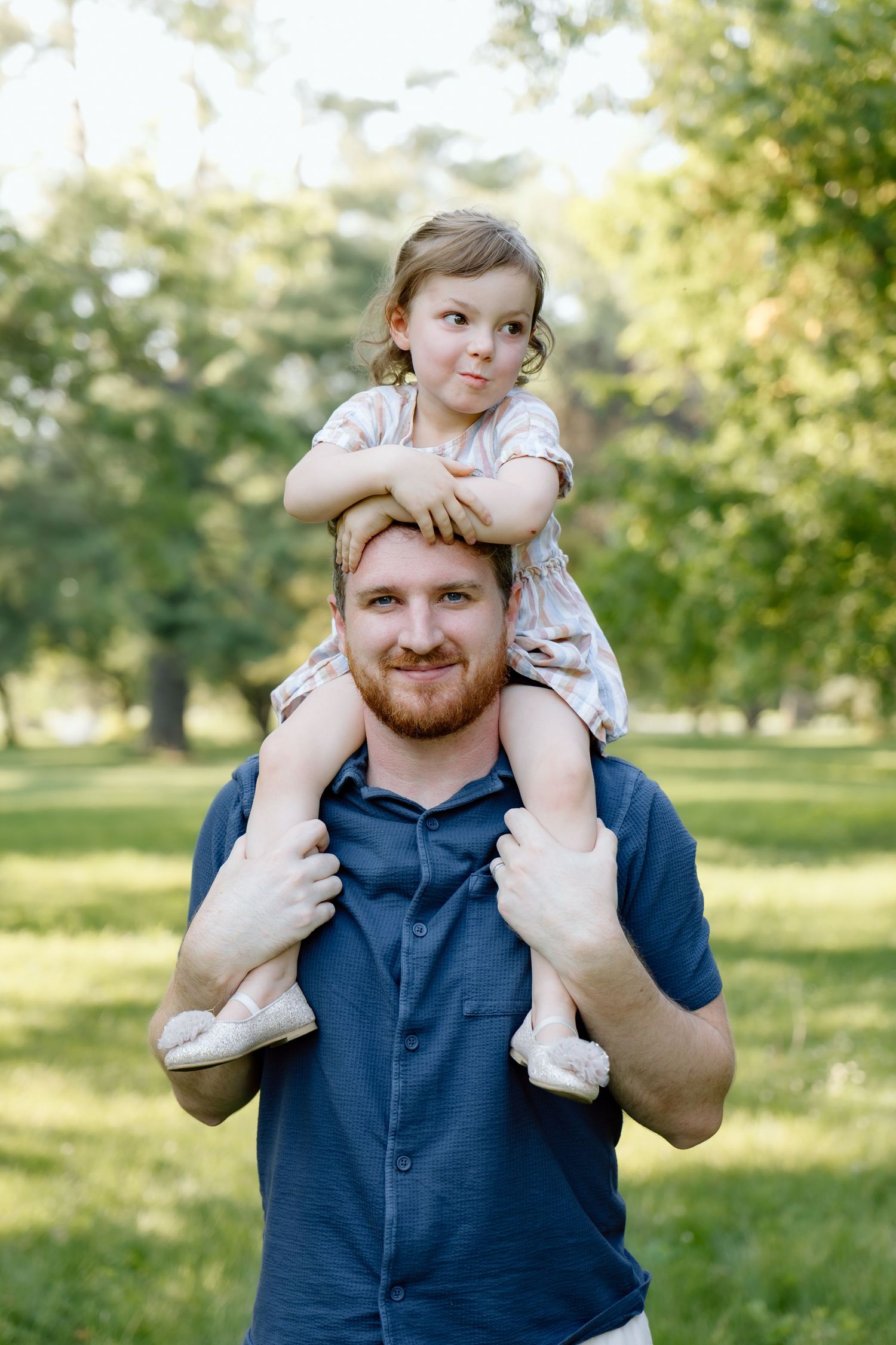 Smiling red-bearded father carrying his toddler daughter on his shoulders in a sunny park.