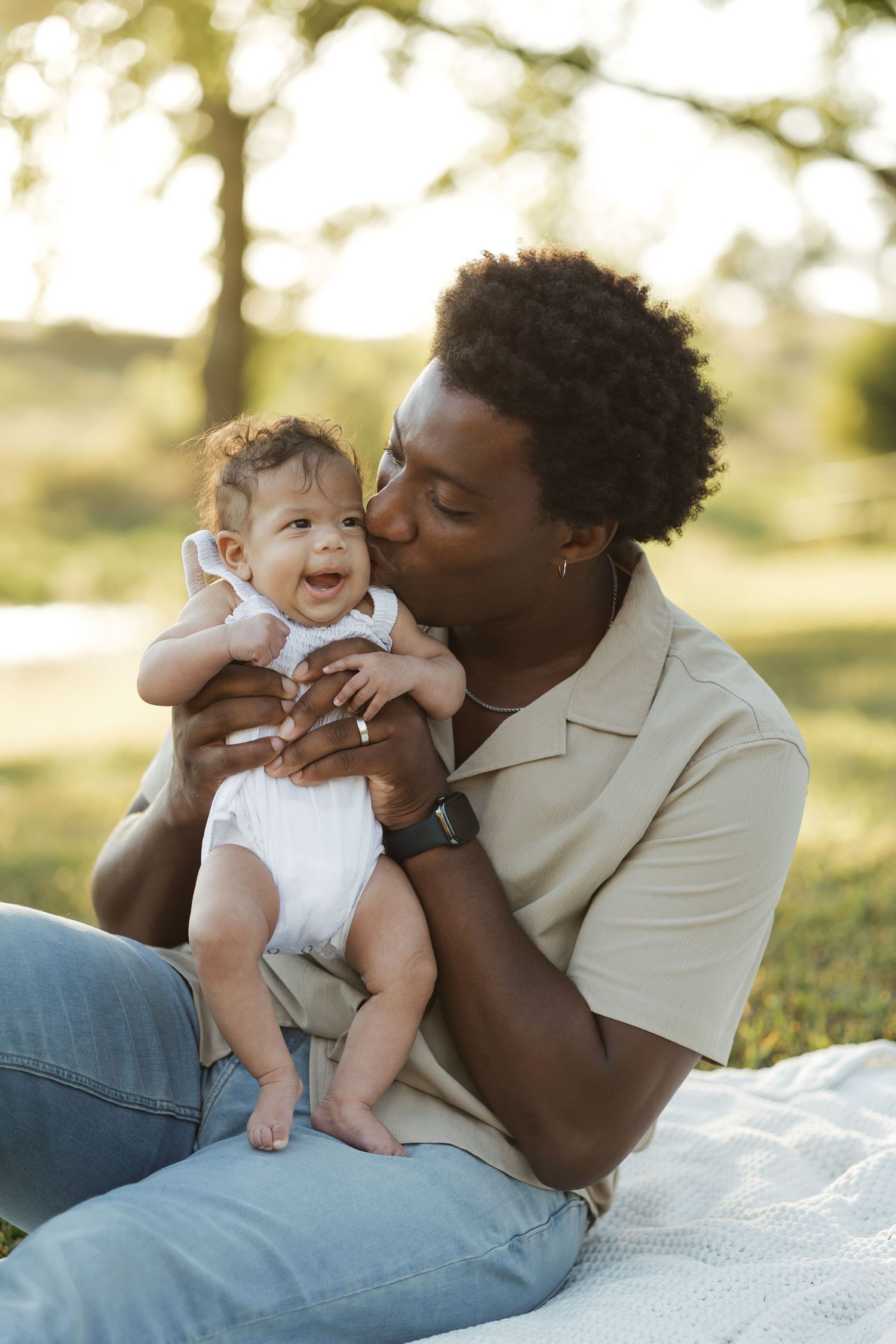 Father kissing laughing baby while sitting on blanket in sunny park.