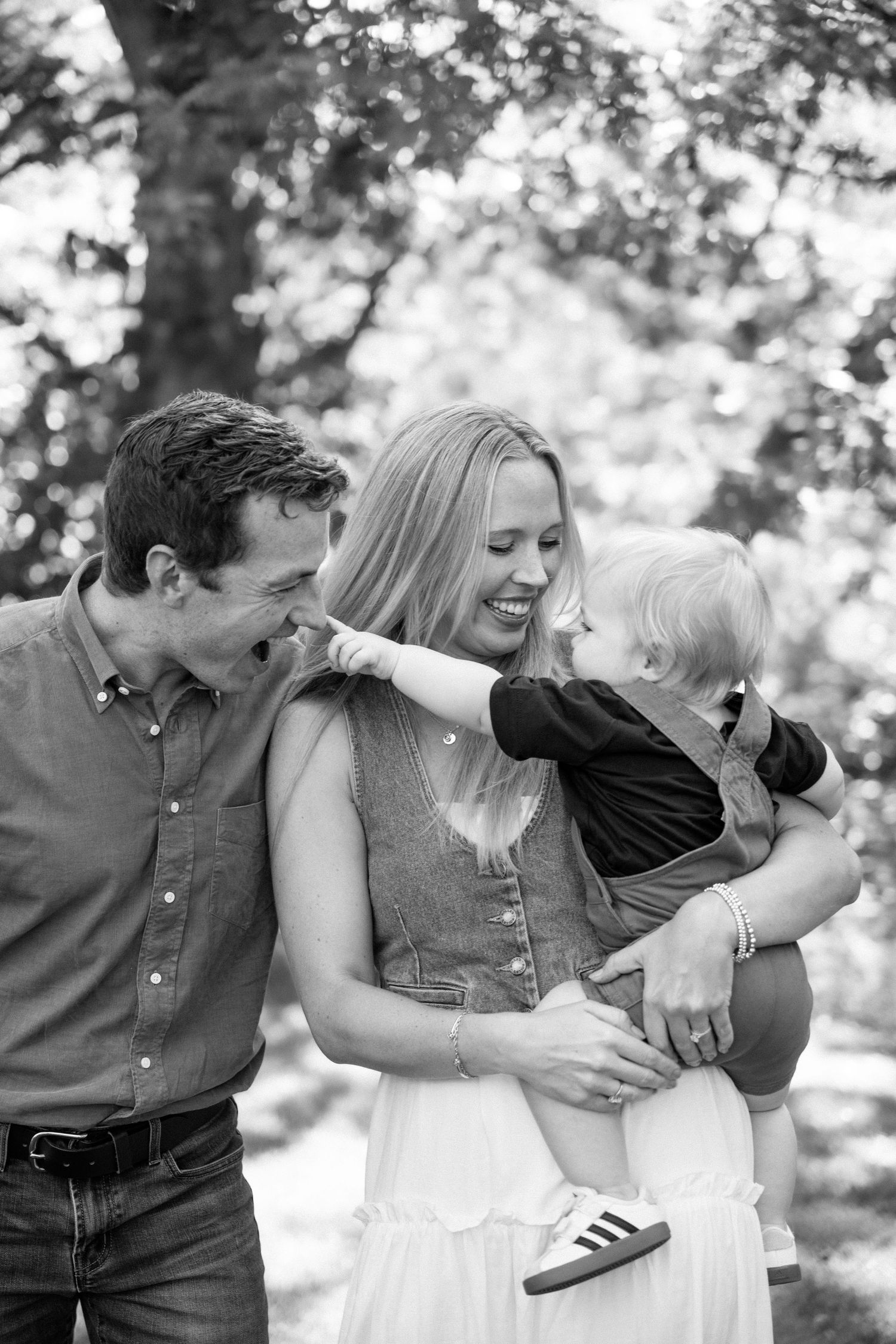 Black and white family portrait of parents laughing as toddler reaches toward mother's face outdoors.