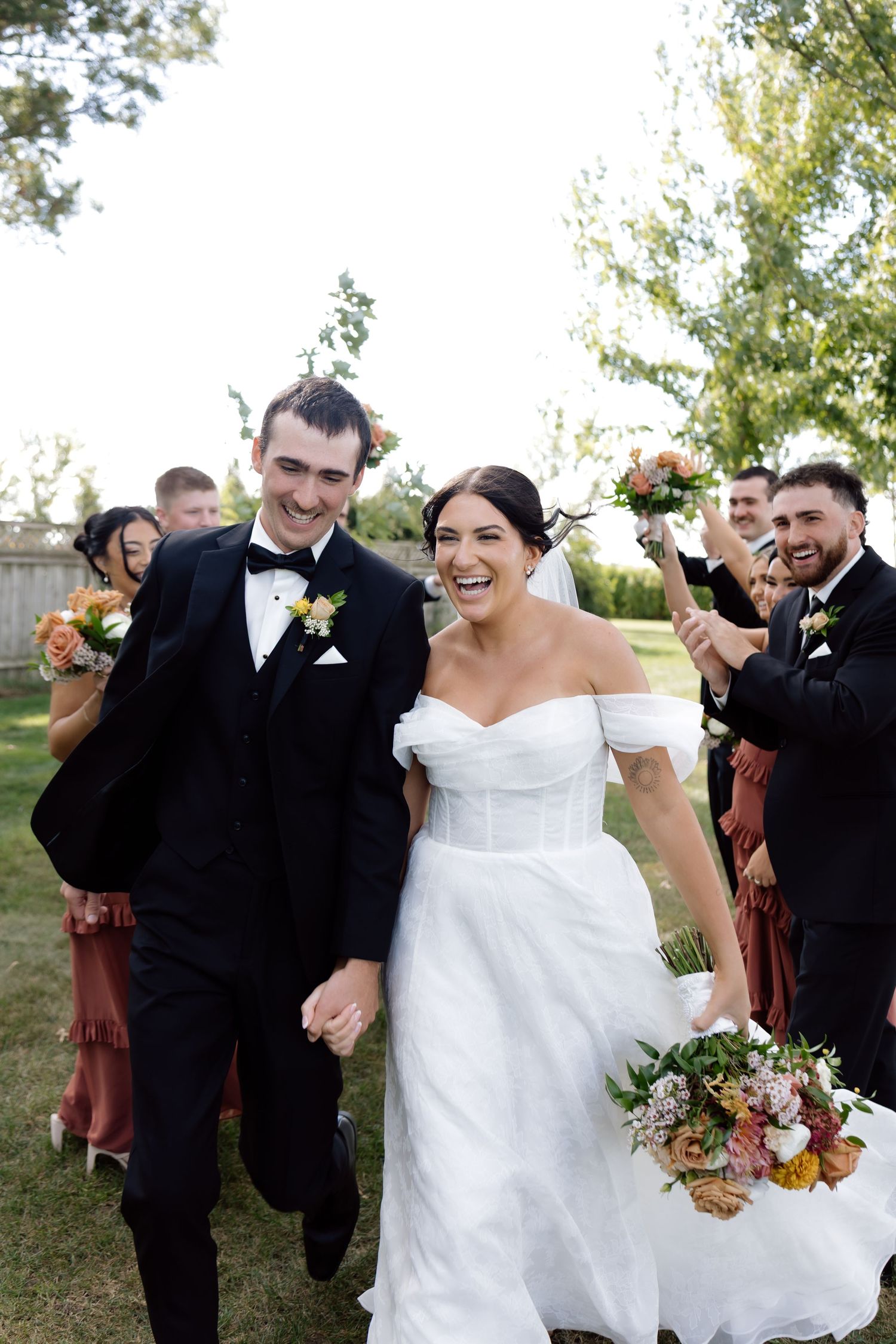 Laughing bride and groom walk through cheering wedding party in outdoor celebration.