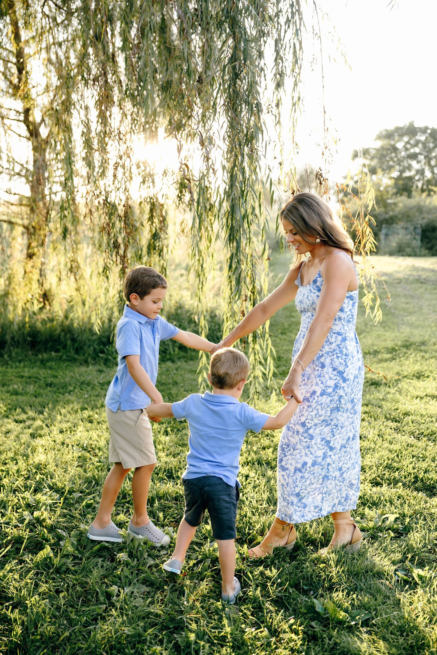 Mother in floral dress holding hands with two young sons under a weeping willow tree at sunset.