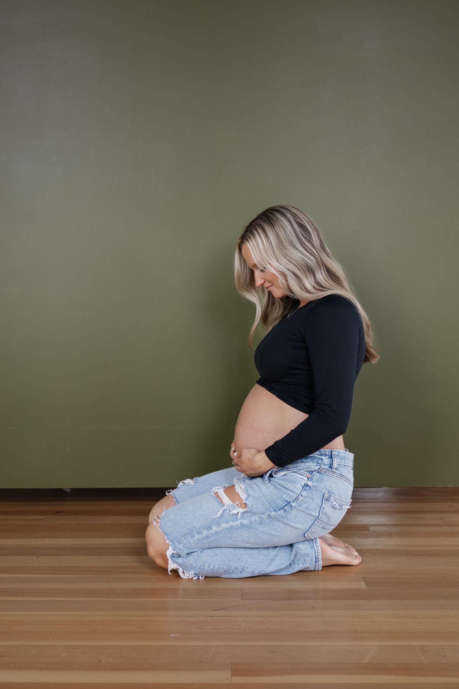 Pregnant woman kneeling on wooden floor against olive green wall, cradling her bare baby bump tenderly.