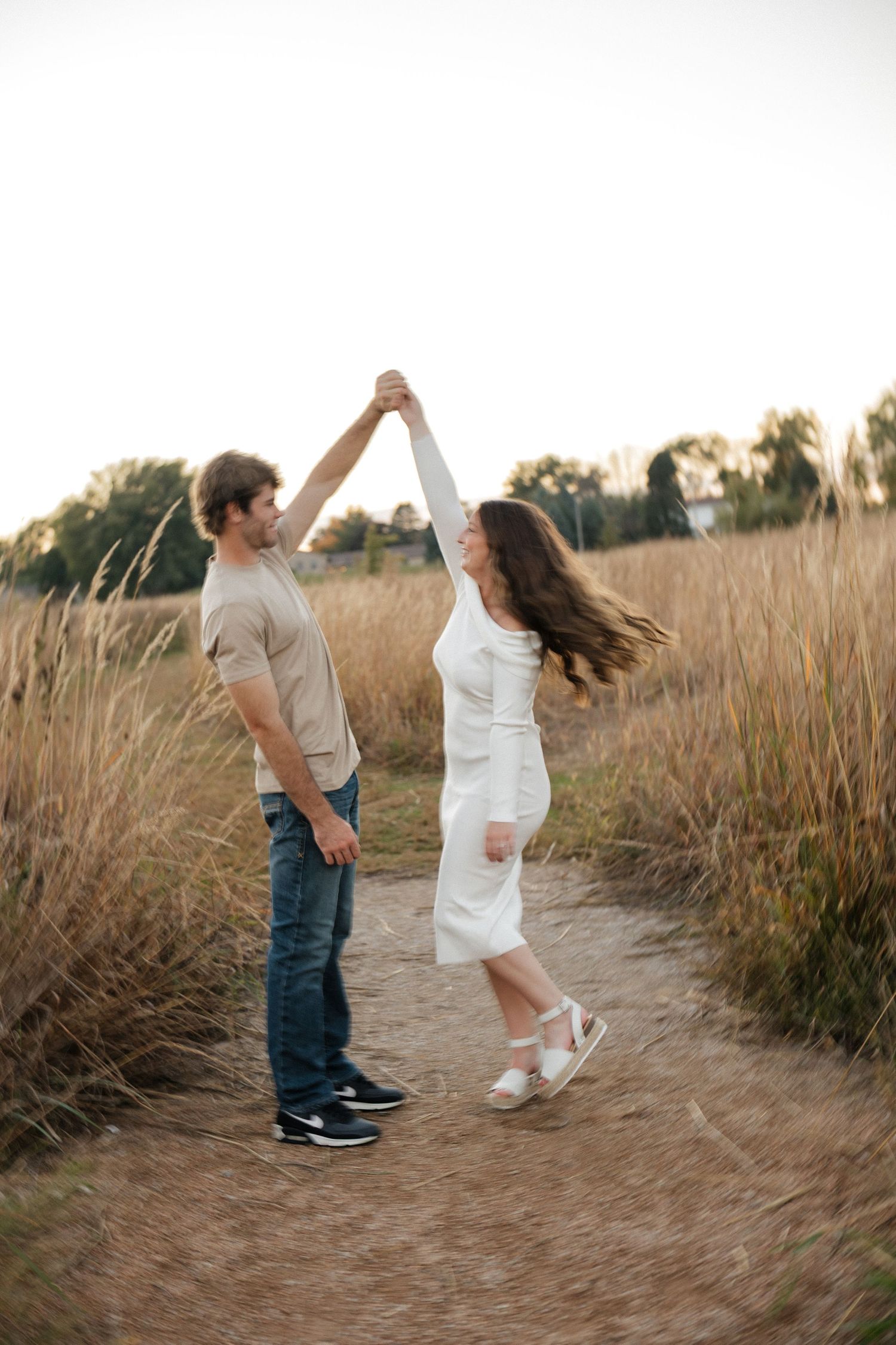 Couple dancing joyfully on a dirt path in a golden field at sunset, woman in flowing white dress.