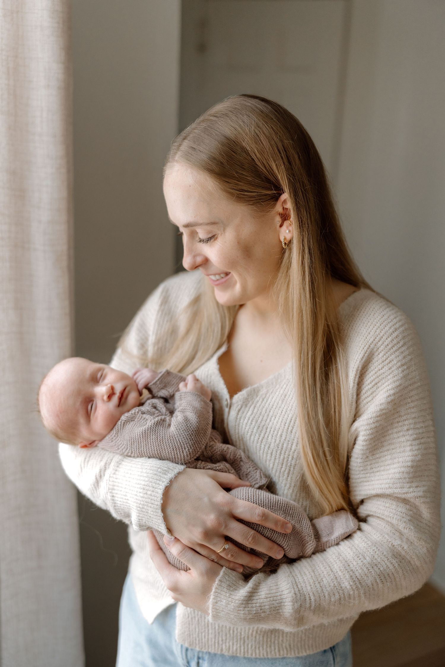 Smiling blonde mother in beige sweater lovingly holds her newborn baby near a bright window.