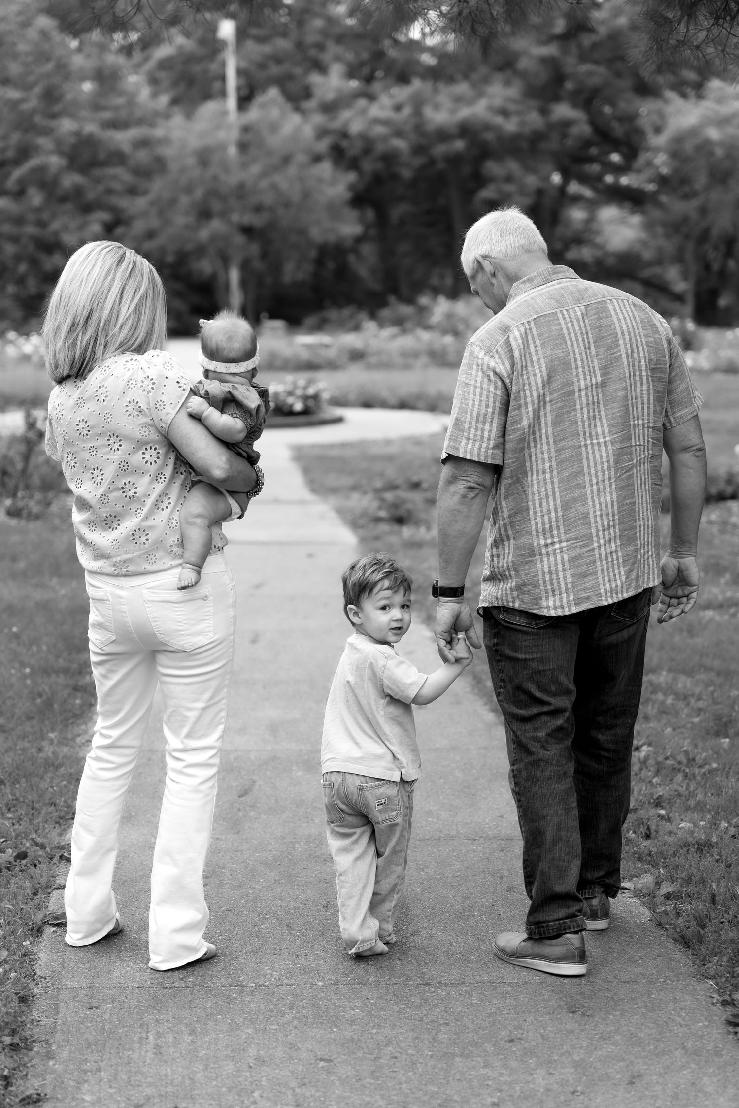 Black and white photo of grandparents walking with toddler and baby on a park path.