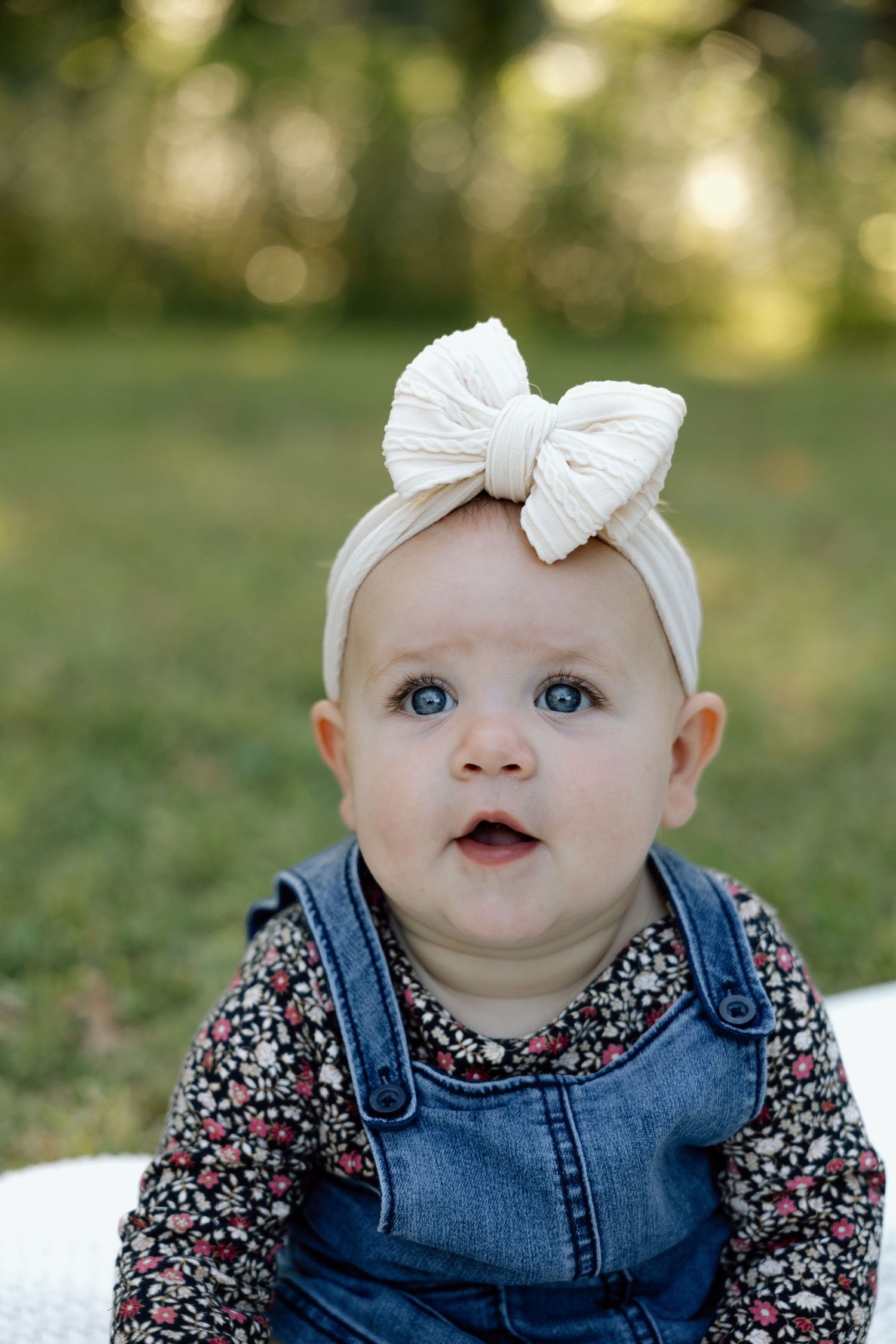 Adorable blue-eyed baby girl wearing a large cream bow headband sitting outdoors in a park.