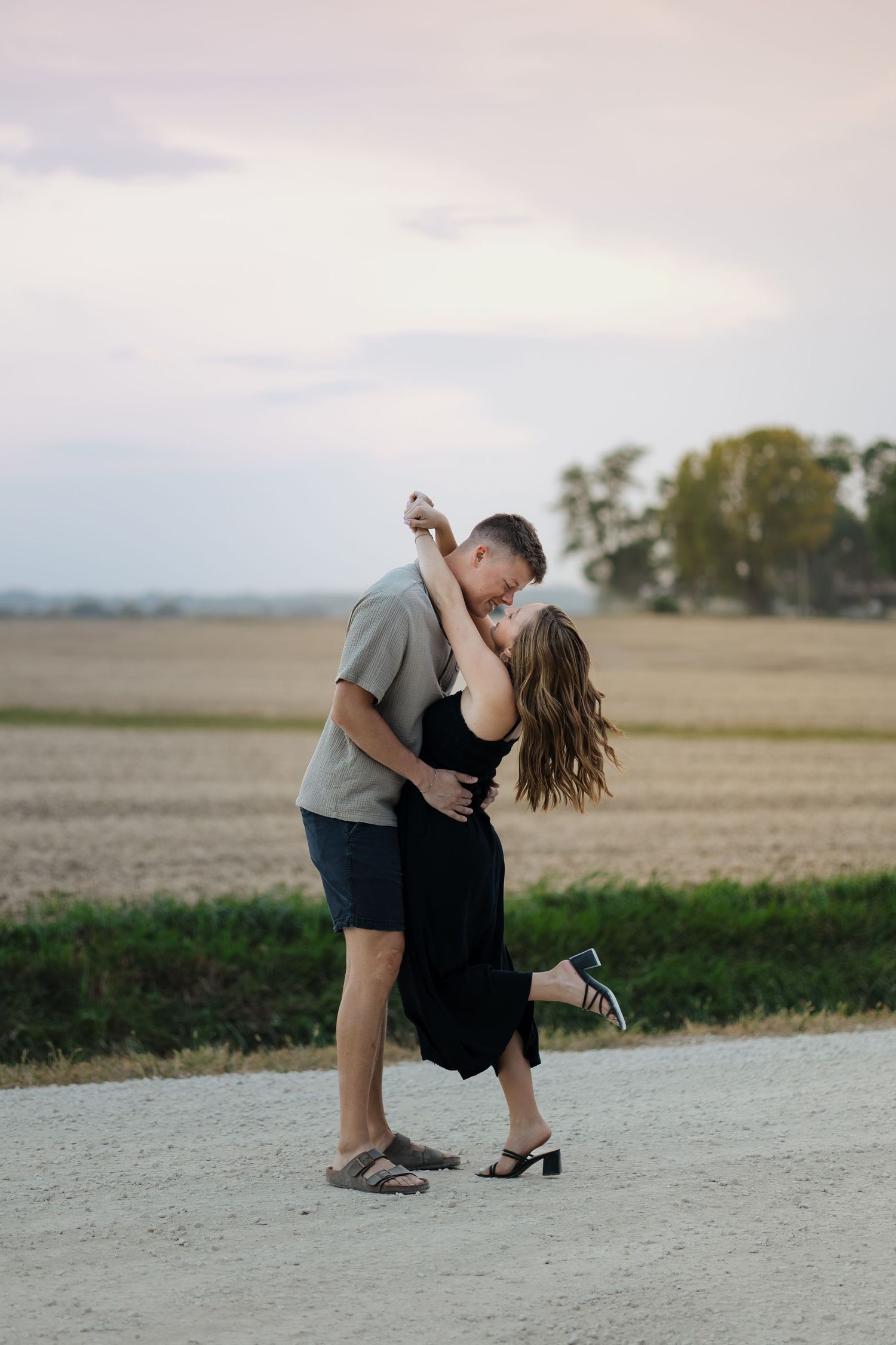 Couple sharing romantic dip kiss on gravel path with flat farm fields at sunset.