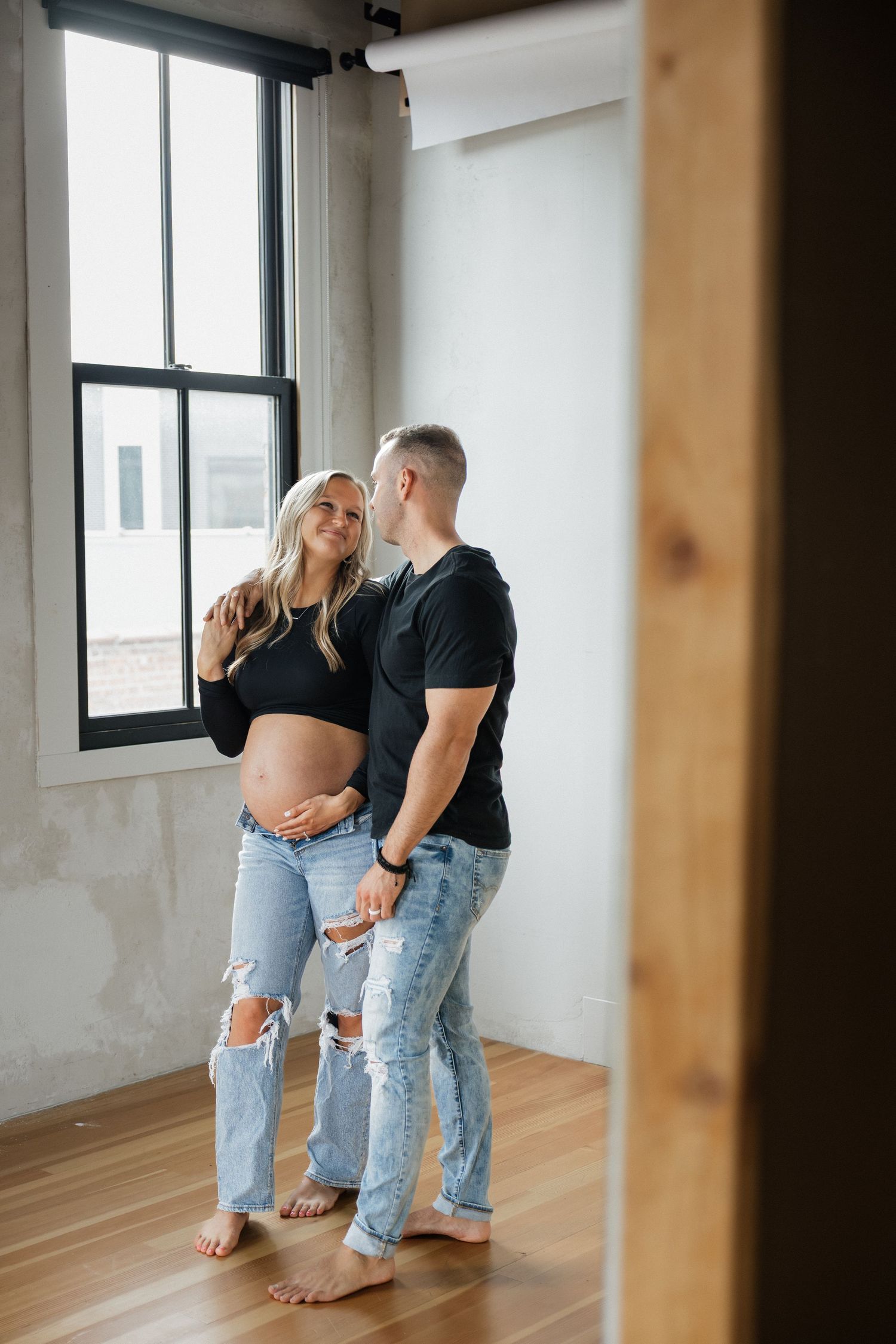 Pregnant woman and partner standing by a large window, sharing an intimate moment while cradling her baby bump.