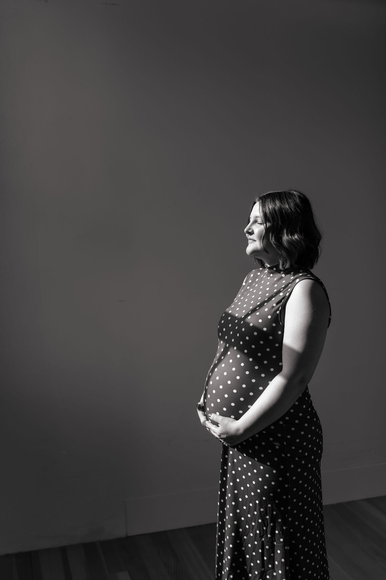 Black and white portrait of a smiling pregnant woman in a polka dot dress cradling her baby bump.