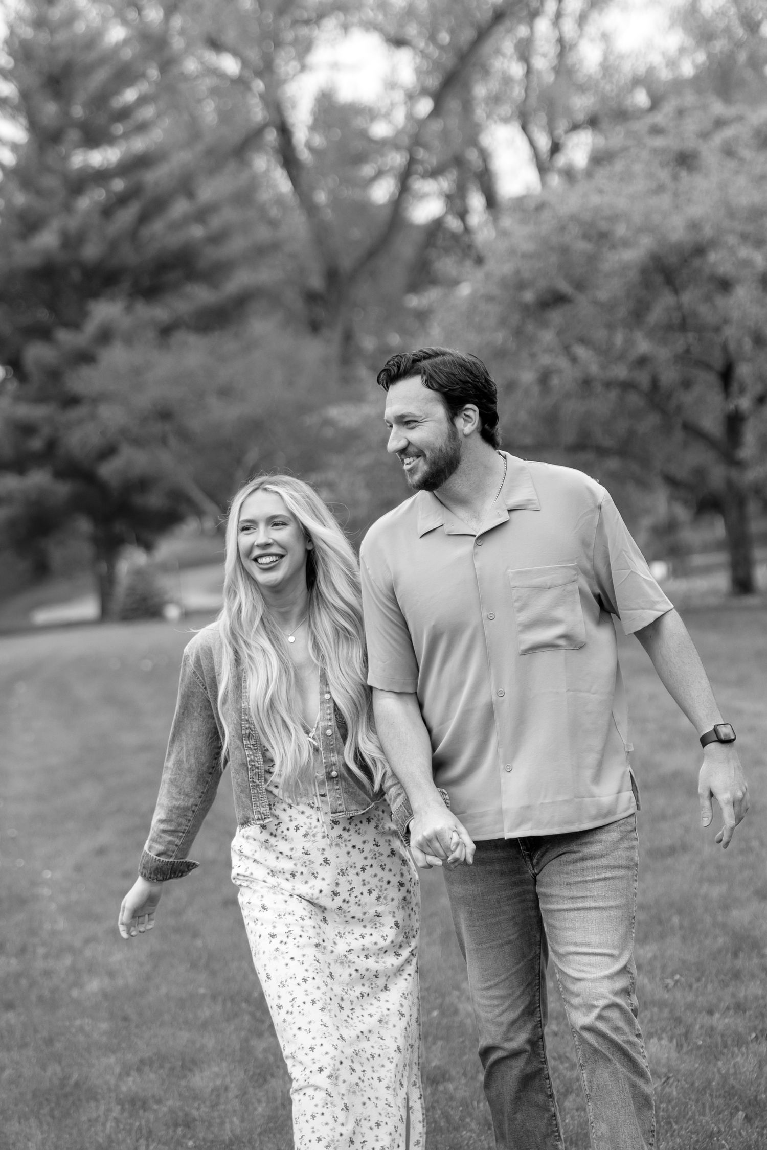 A smiling couple holds hands while walking through a lush green park in this black and white photo.