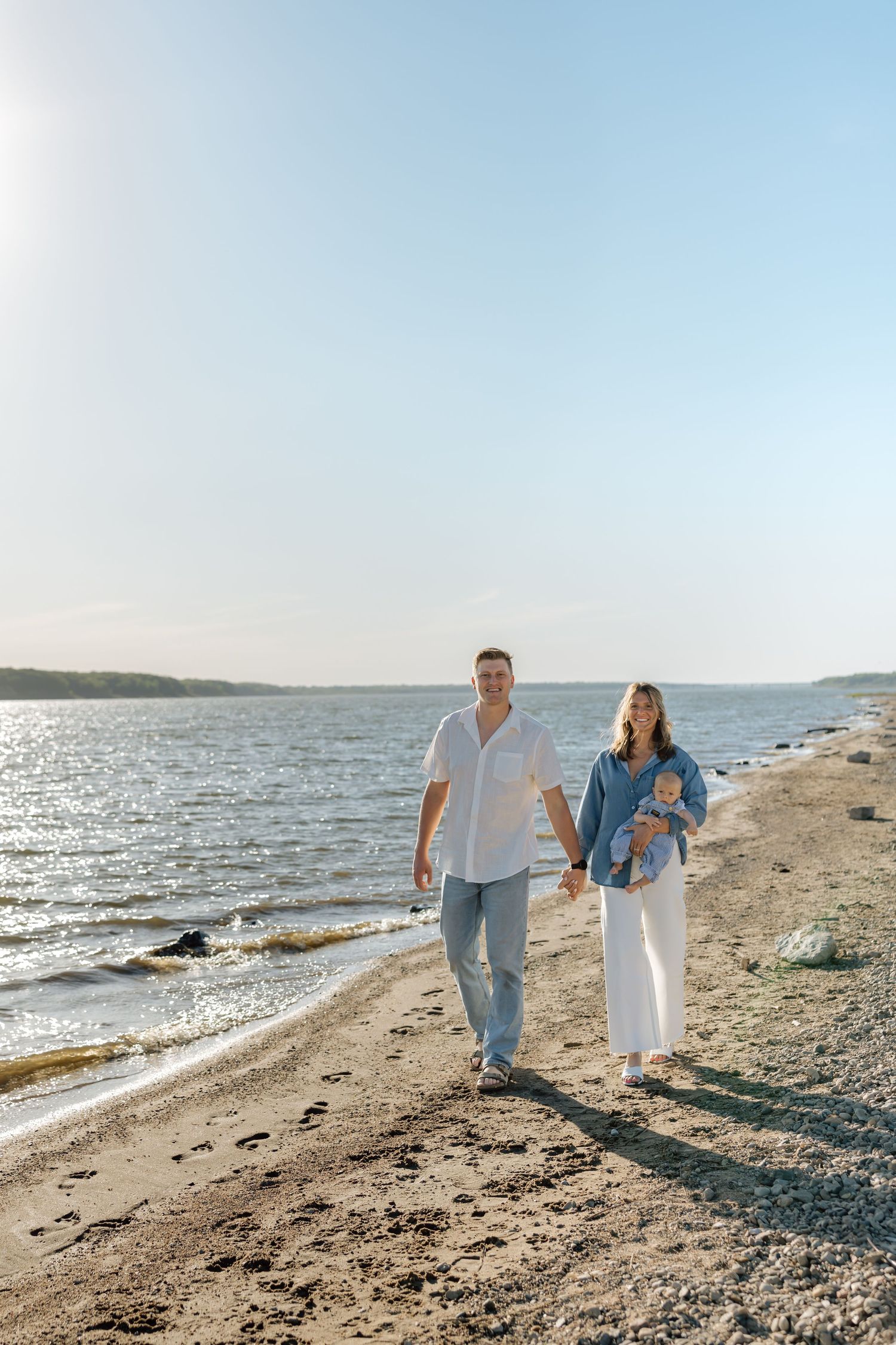 A couple walks hand-in-hand along a sunny beach shoreline, the woman carrying a baby while wearing casual summer clothes.