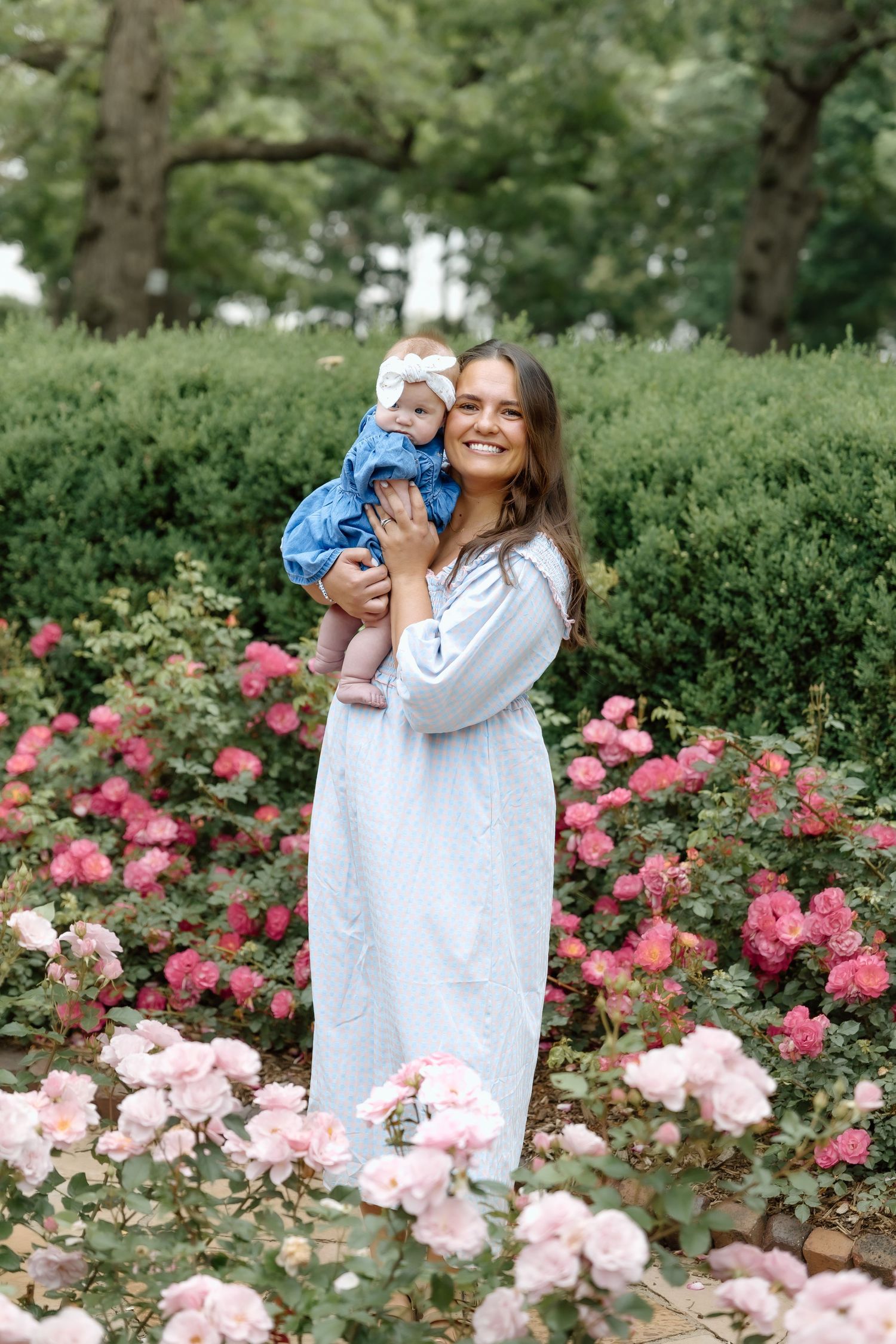 Radiant mother in white dress holds smiling baby girl in blue outfit among blooming pink roses.
