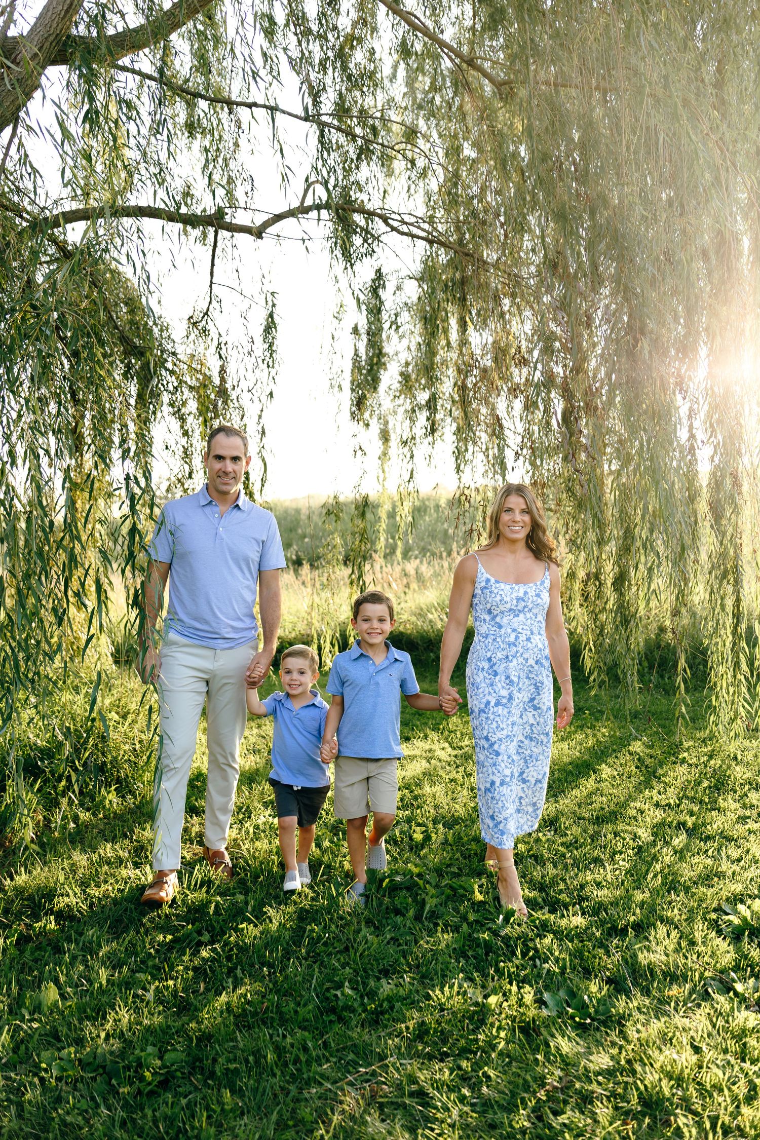 Family of four holding hands walking under willow tree in sunny field.